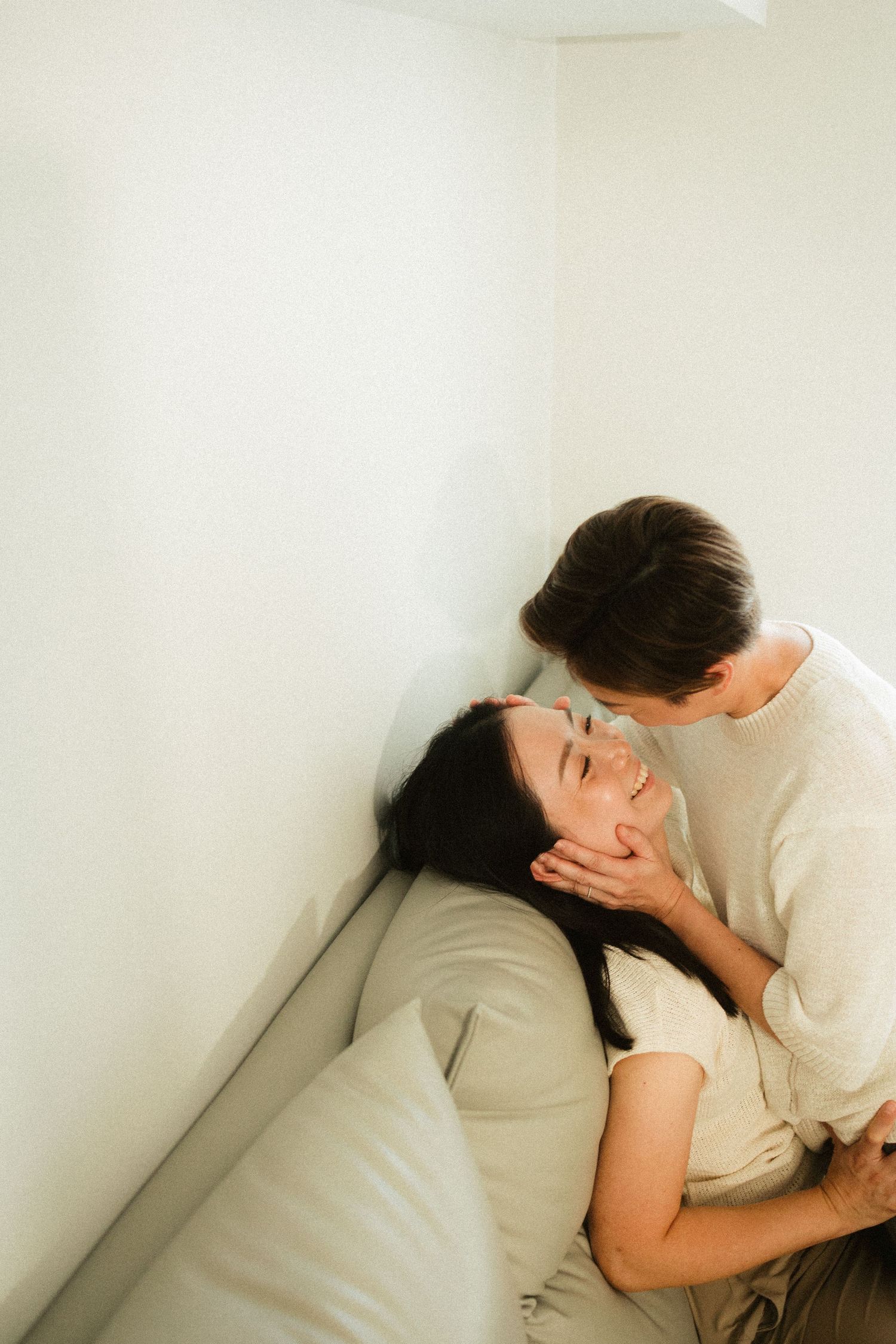 Asian couple sharing a tender moment with one leaning down to kiss the other on sofa.