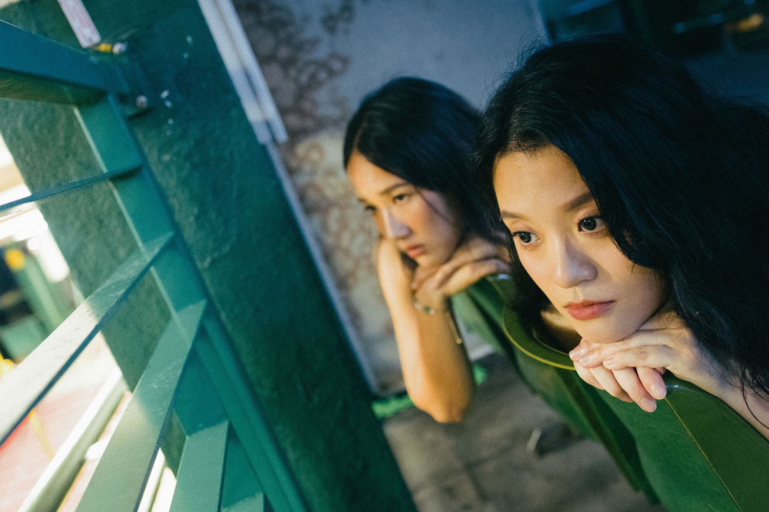 Two young Asian women rest their chins on hands while gazing through green railings.