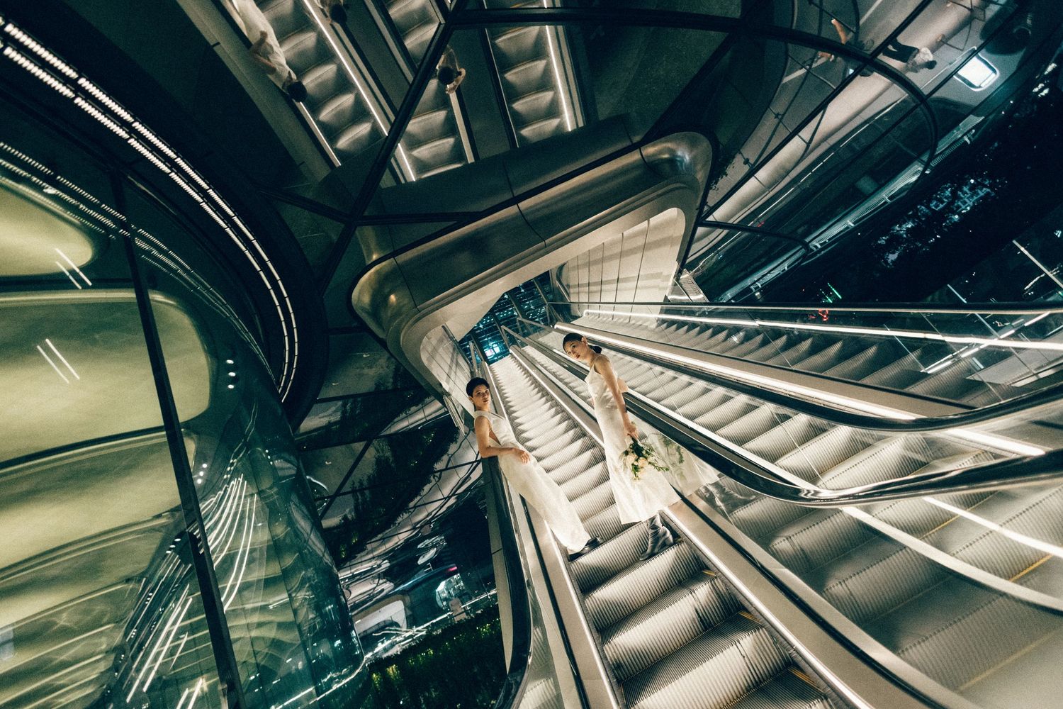 Aerial view of two people in white outfits riding escalators in a modern glass building at night.