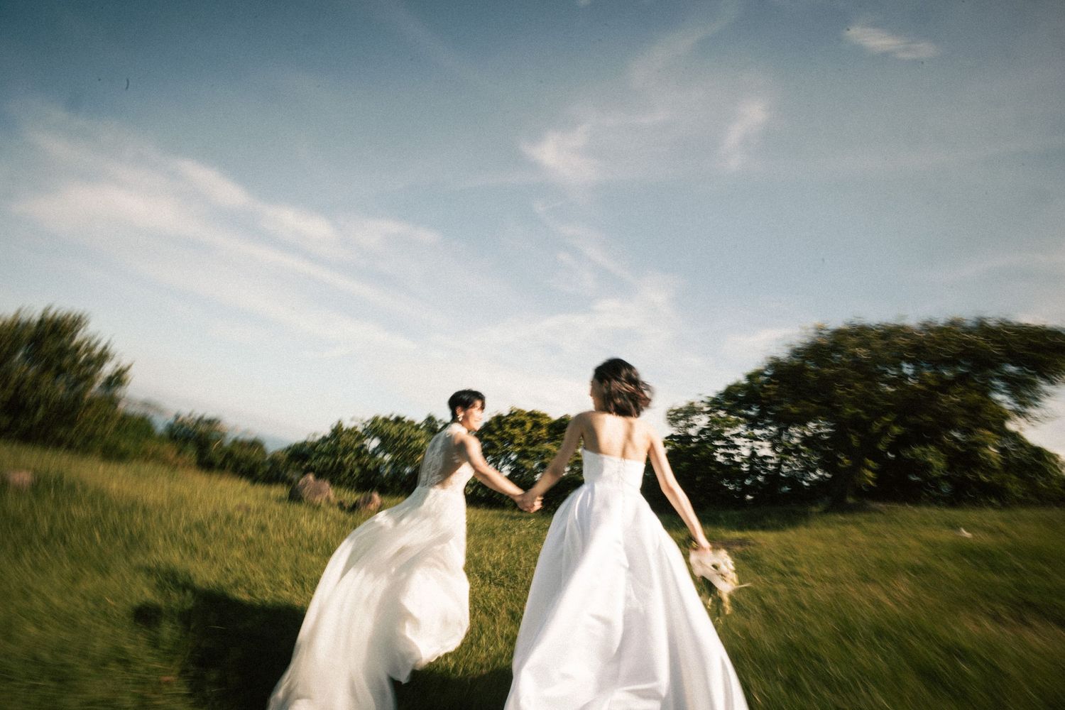 Two brides in flowing white gowns run hand-in-hand across a green meadow under a bright blue sky.