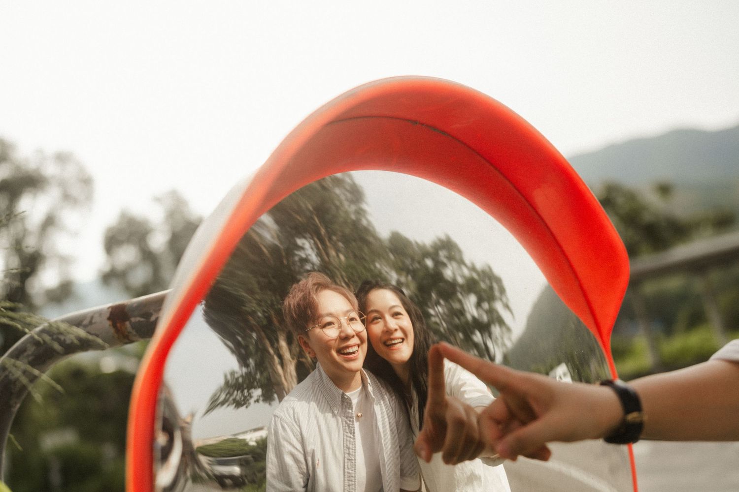 Two smiling women are reflected in a round red-framed mirror outdoors on a sunny day.
