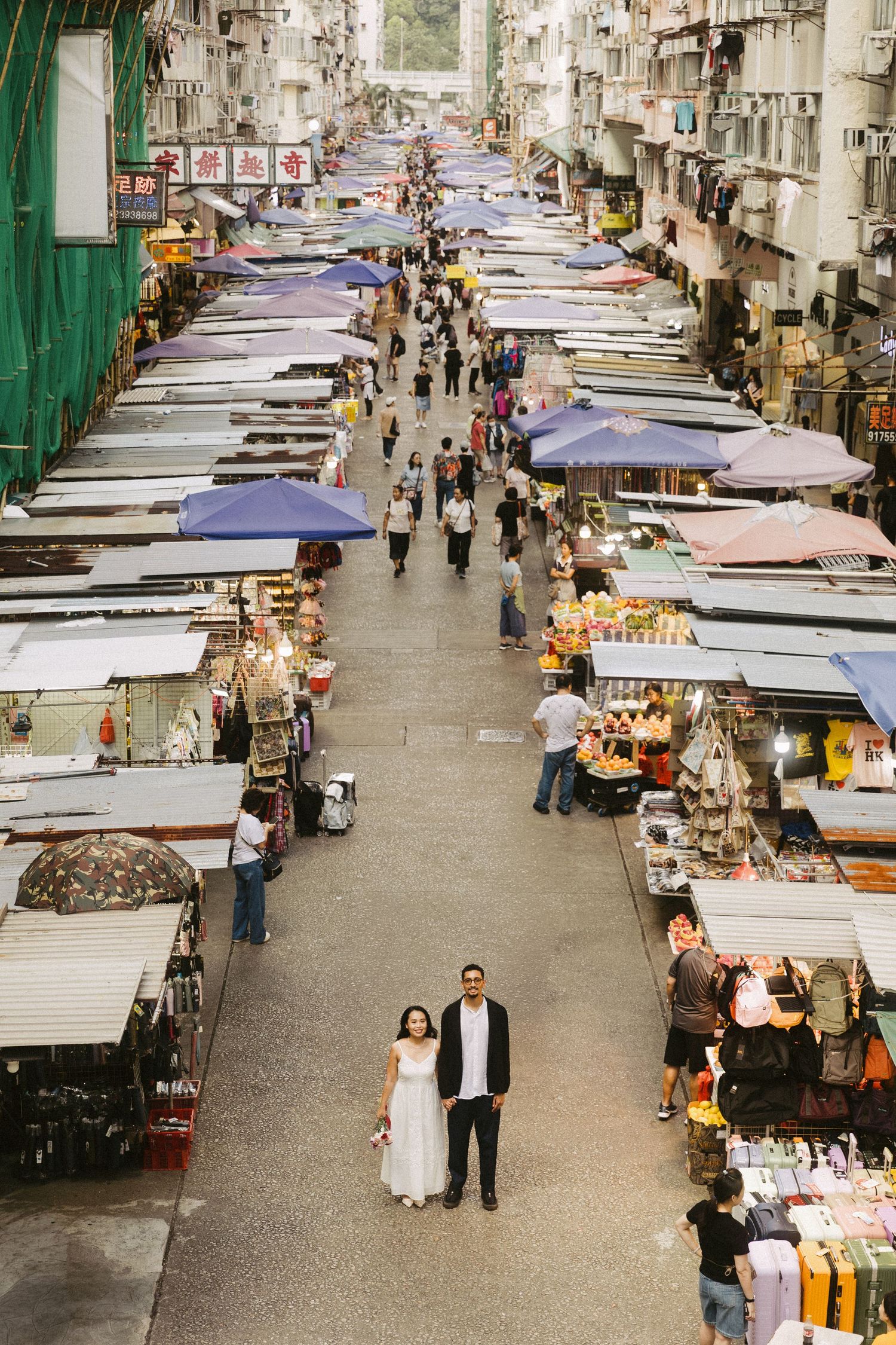 Couple in wedding attire standing in busy Hong Kong street market with colorful vendor stalls and umbrellas.
