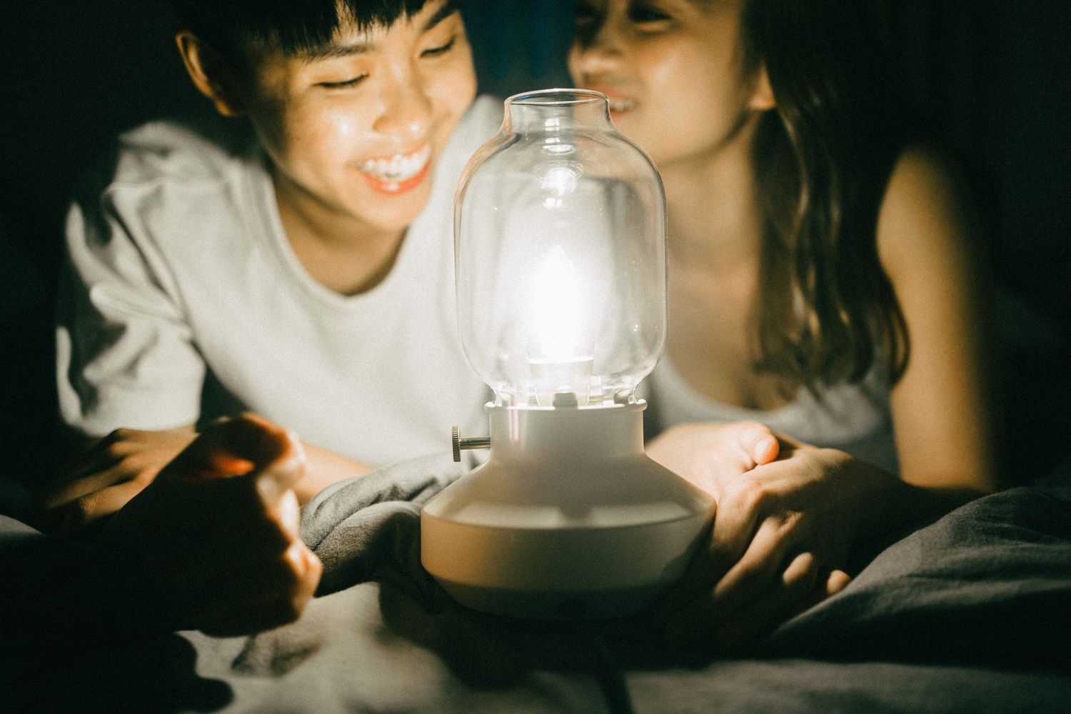 Two children smiling and looking at a glowing lantern lamp together in the dark.
