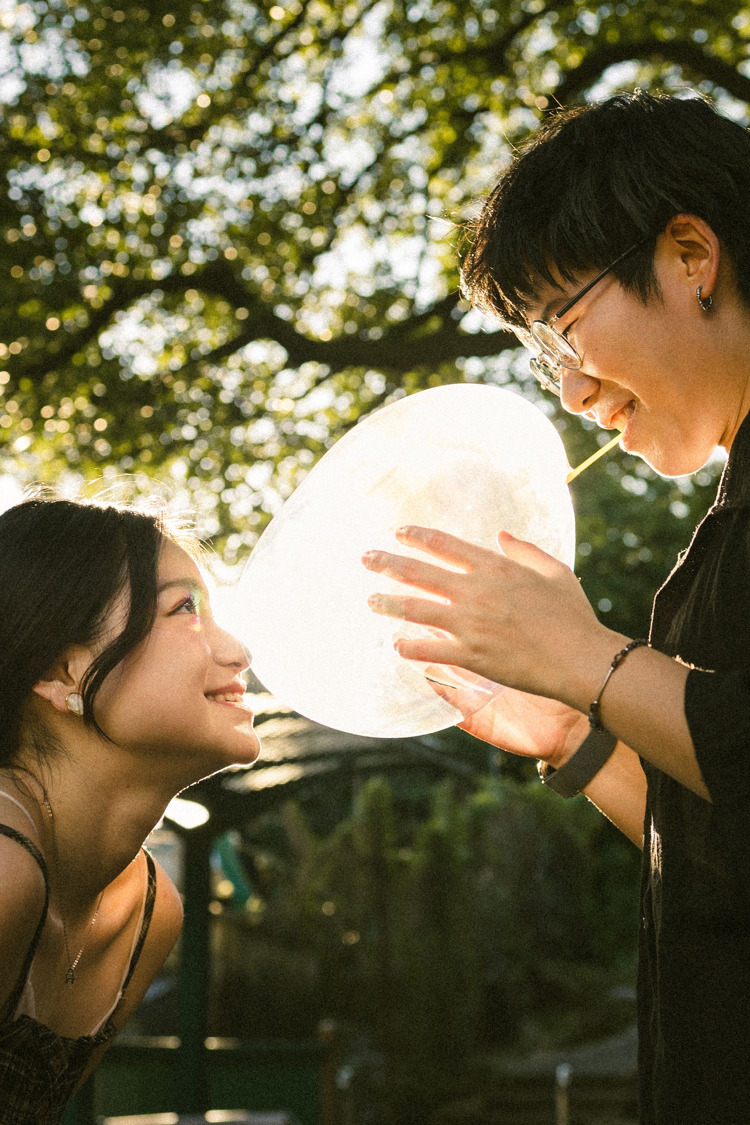 Smiling couple holding a glowing heart-shaped balloon together in a sunlit park setting.