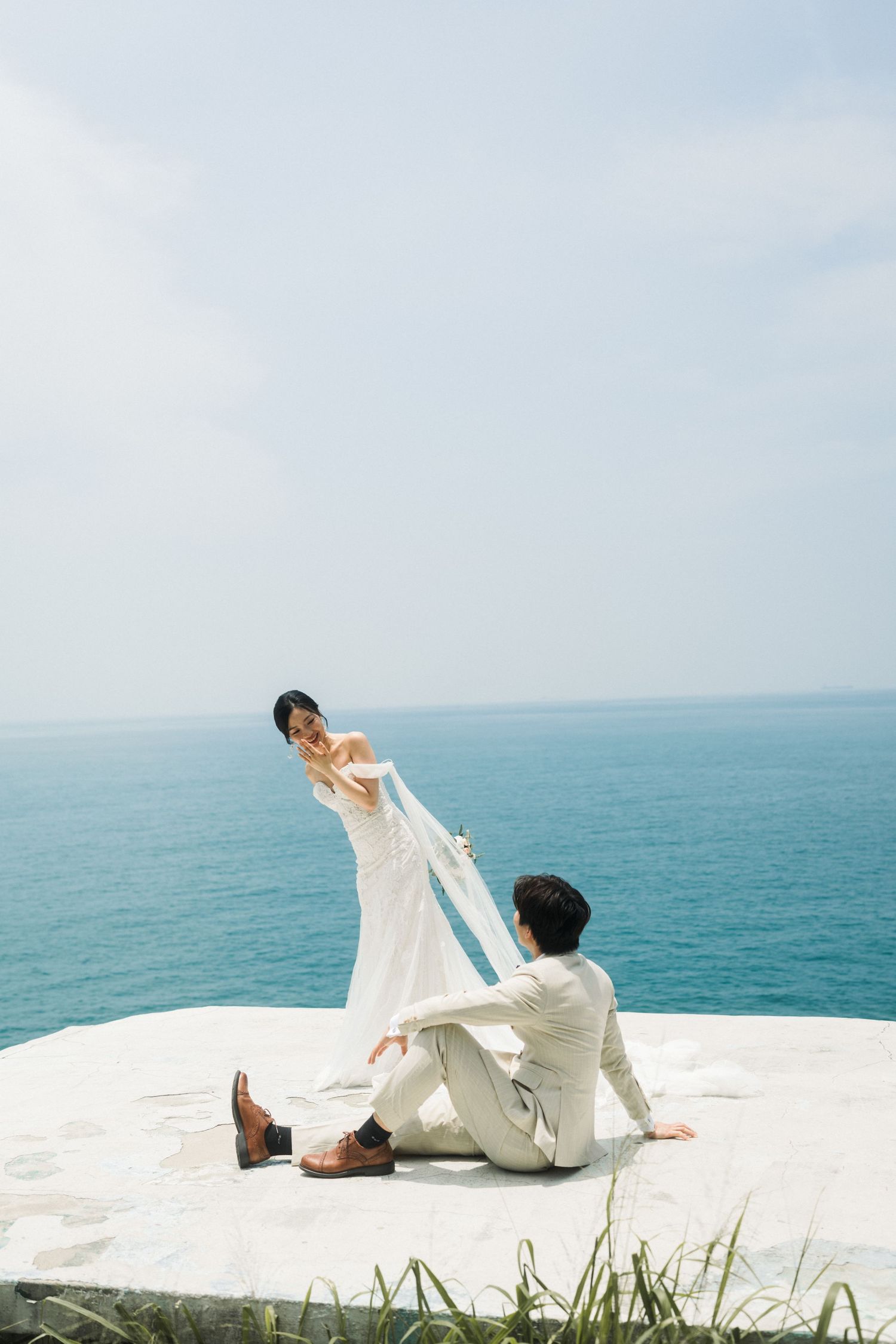 Bride in white gown holds veil while groom sits on seaside platform with ocean in background.