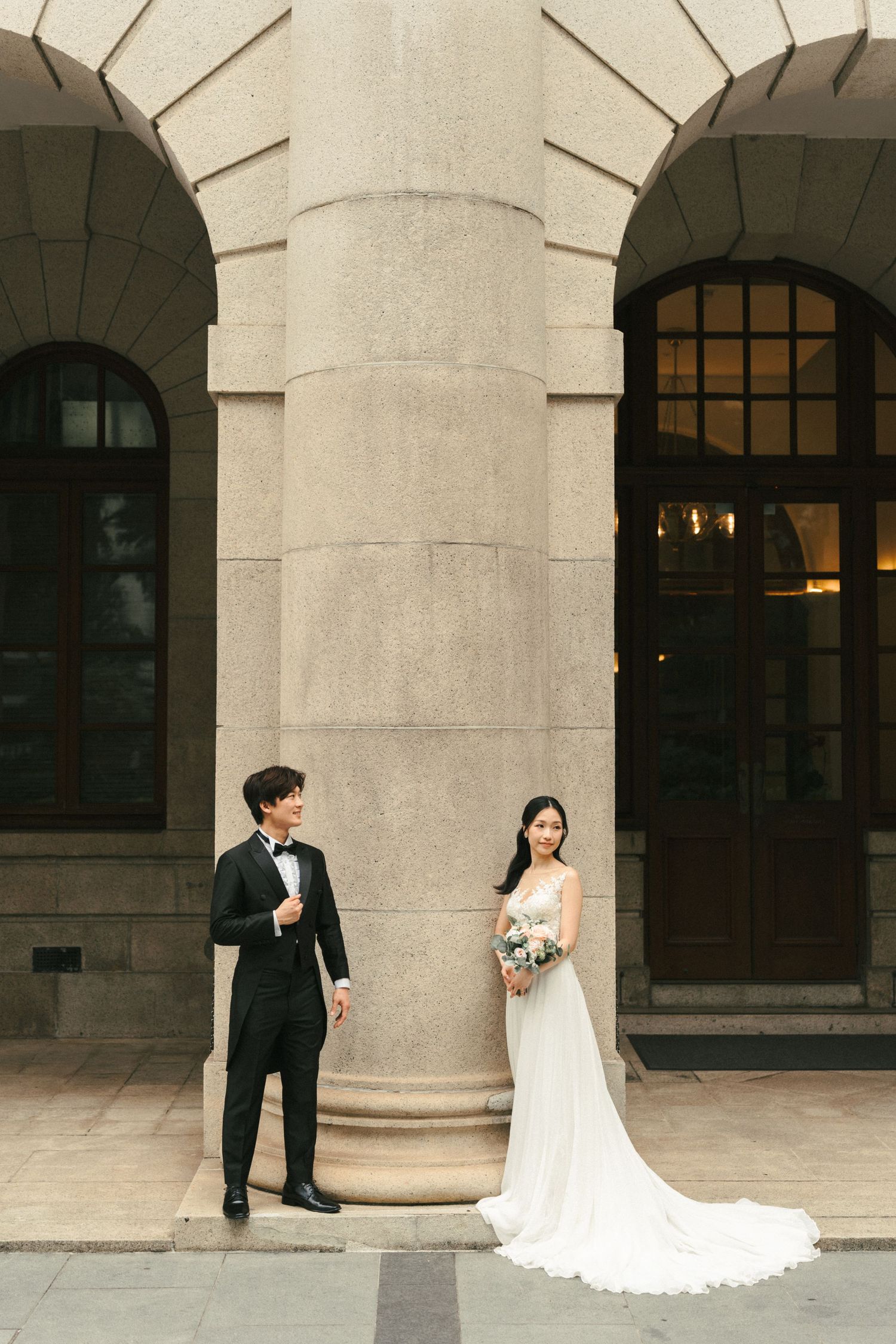 Bride and groom in formal wedding attire posing beside grand stone columns of a classical building.