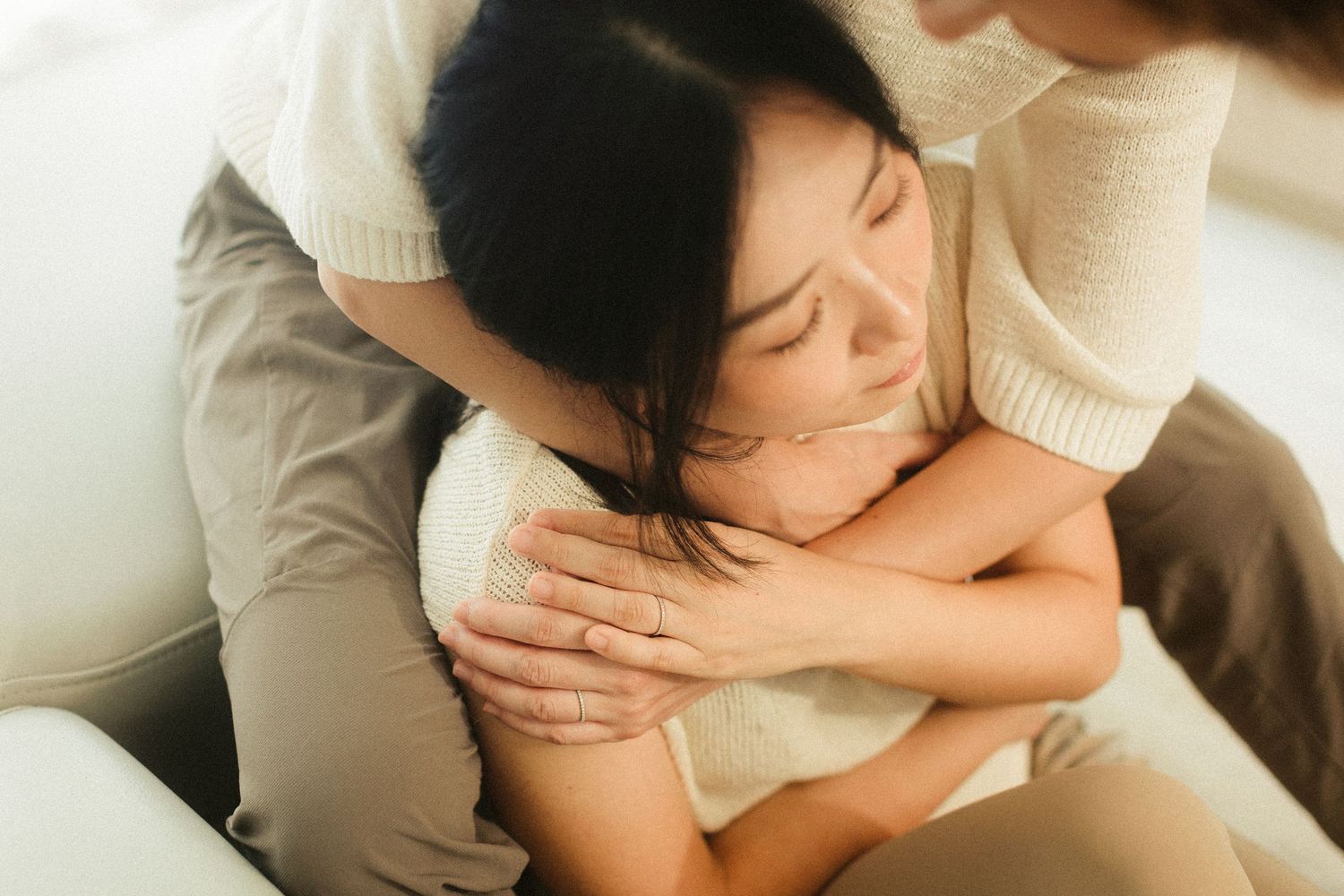 A woman with closed eyes being tenderly hugged from behind while sitting on a sofa.