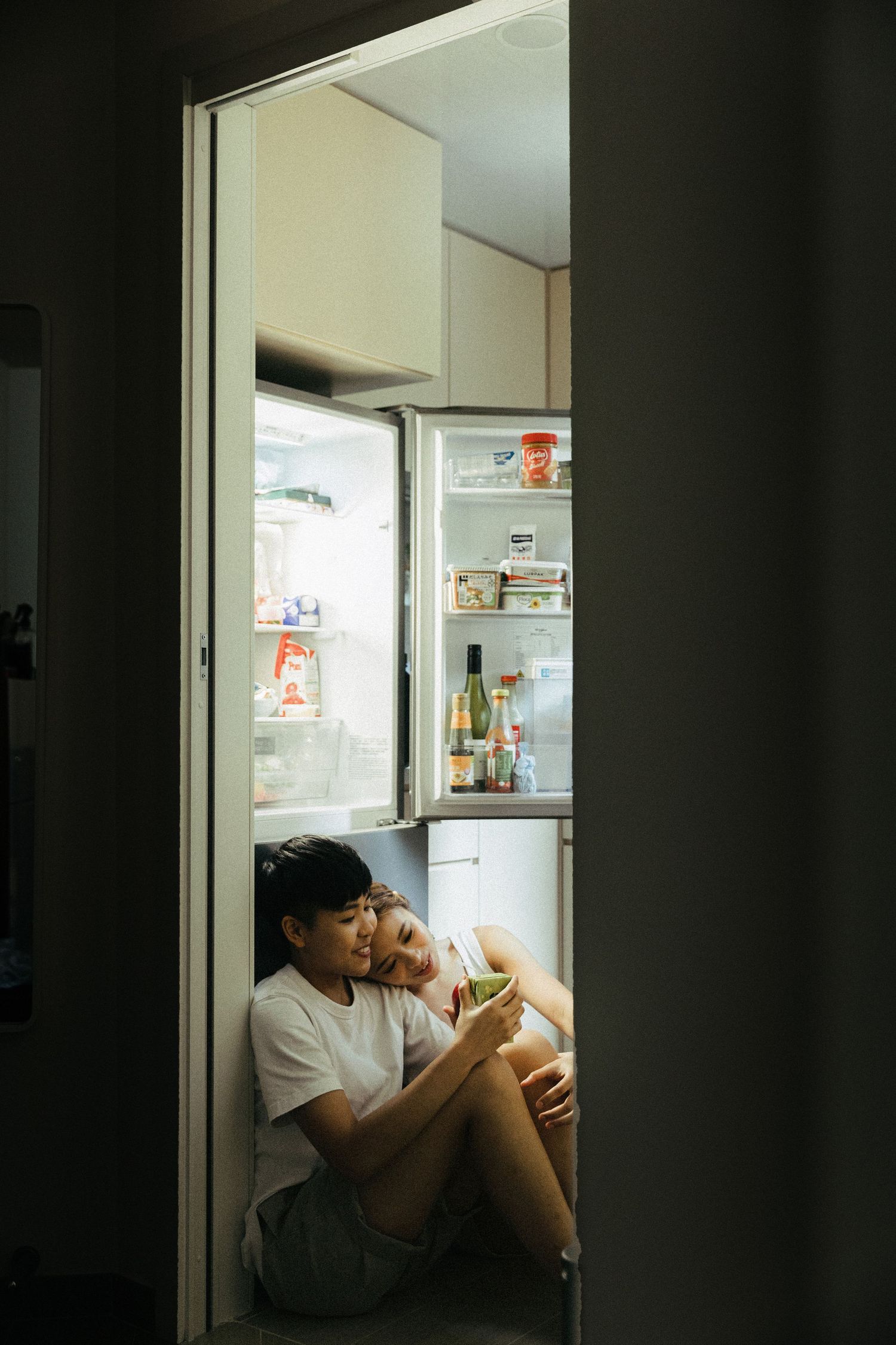 A young Asian couple sitting on the kitchen floor in front of an open refrigerator at night.