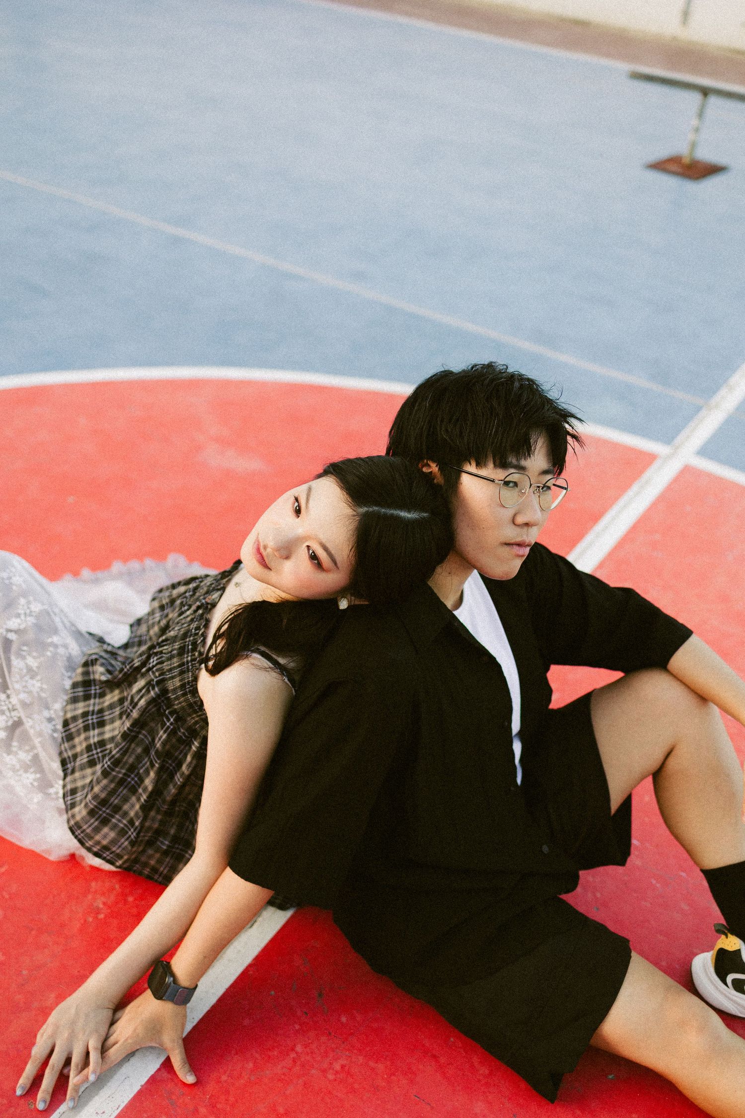 Young couple sitting back-to-back on a red basketball court in casual black outfits.