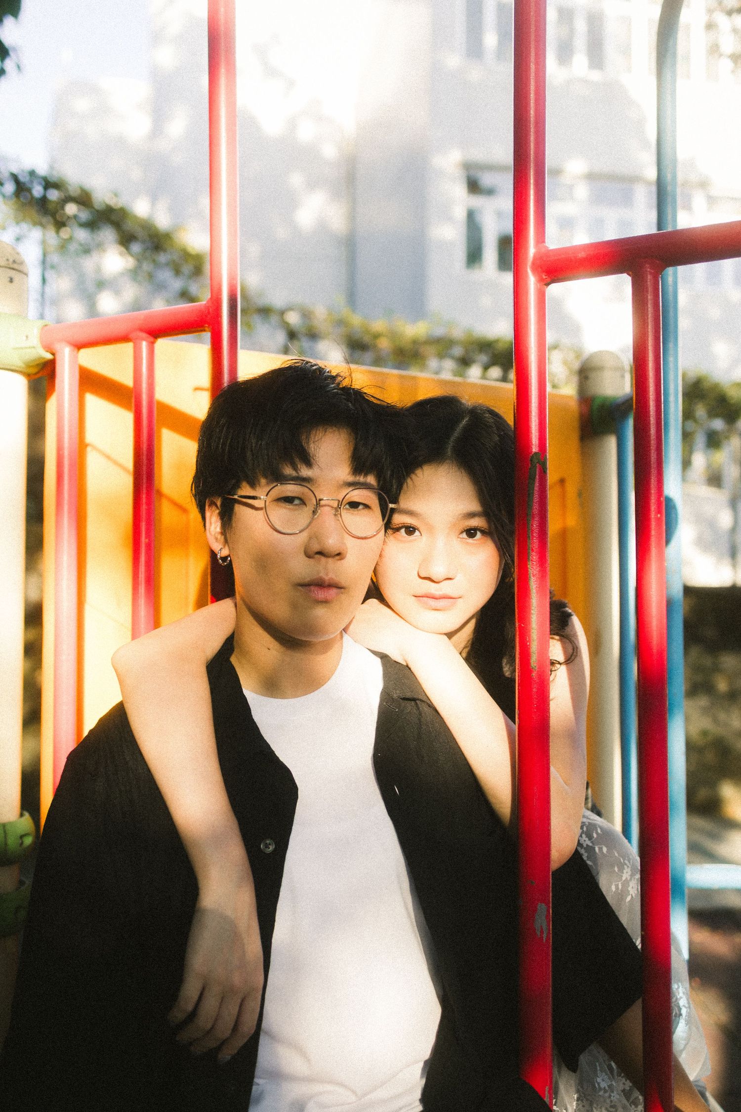 Young Asian couple leaning together against colorful red and yellow playground bars in sunlight.