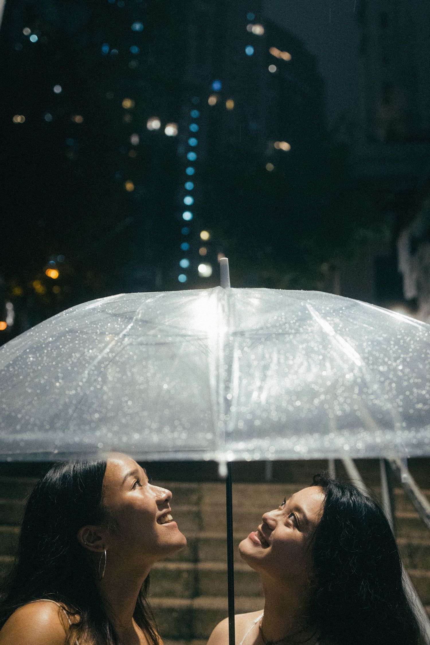 Two young Asian women sharing a clear umbrella in the rain at night.