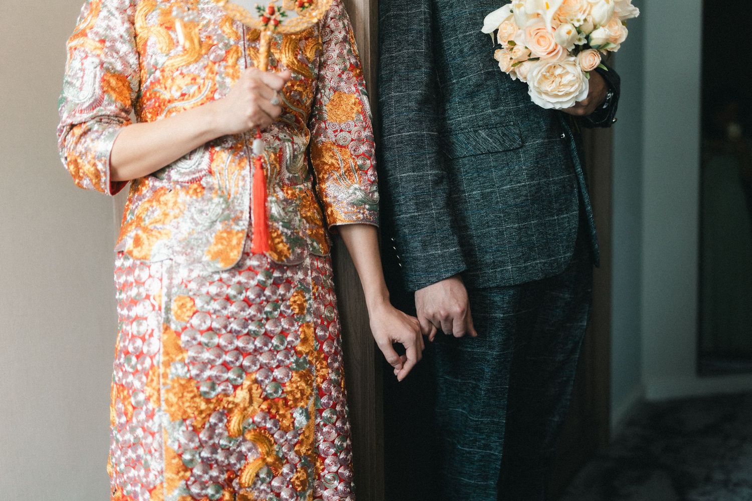 Chinese bride and groom hold hands, bride wearing vibrant embroidered orange qipao holding traditional red tassel fan.