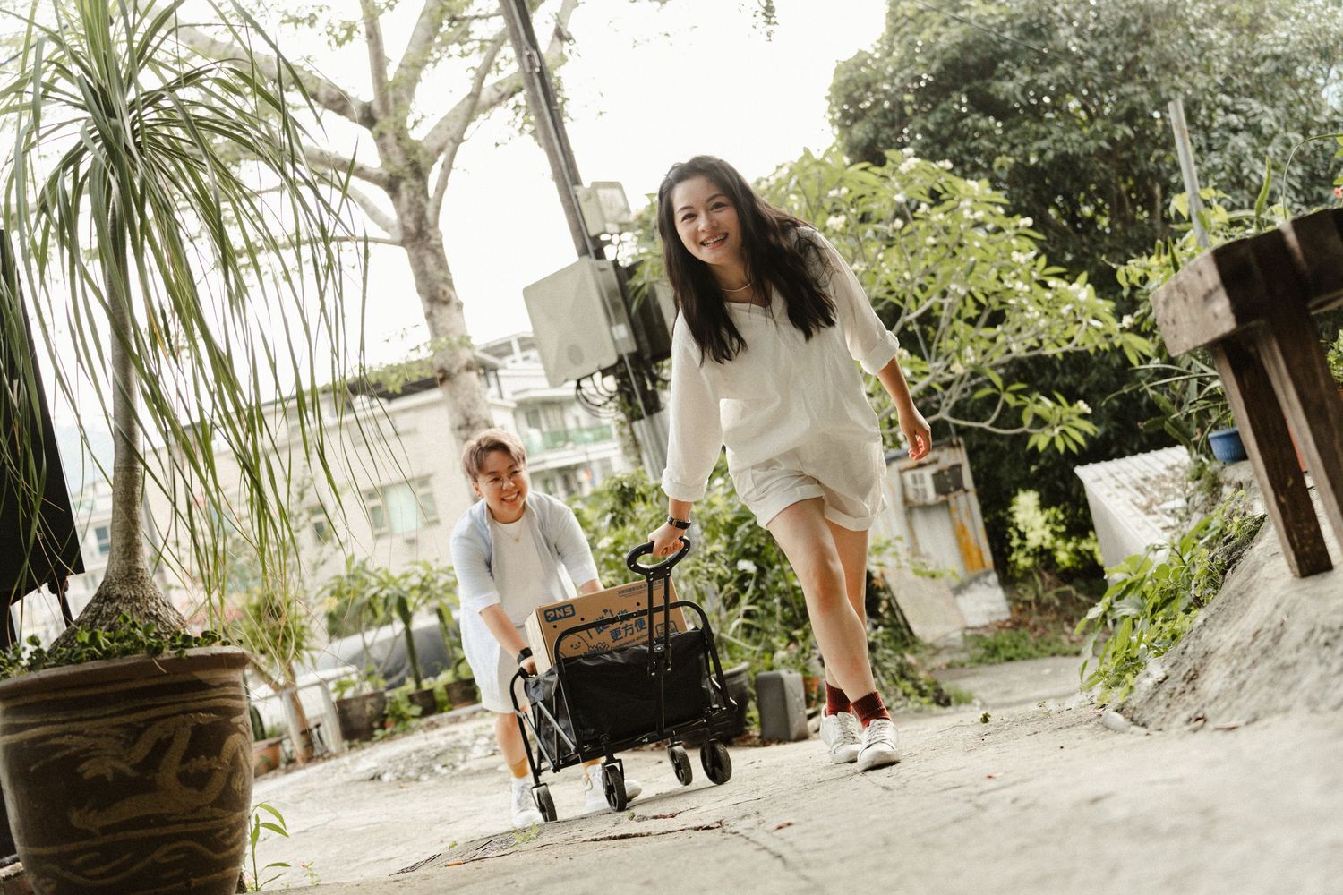 Two women laugh and pull a wagon along a sunny outdoor path surrounded by greenery.