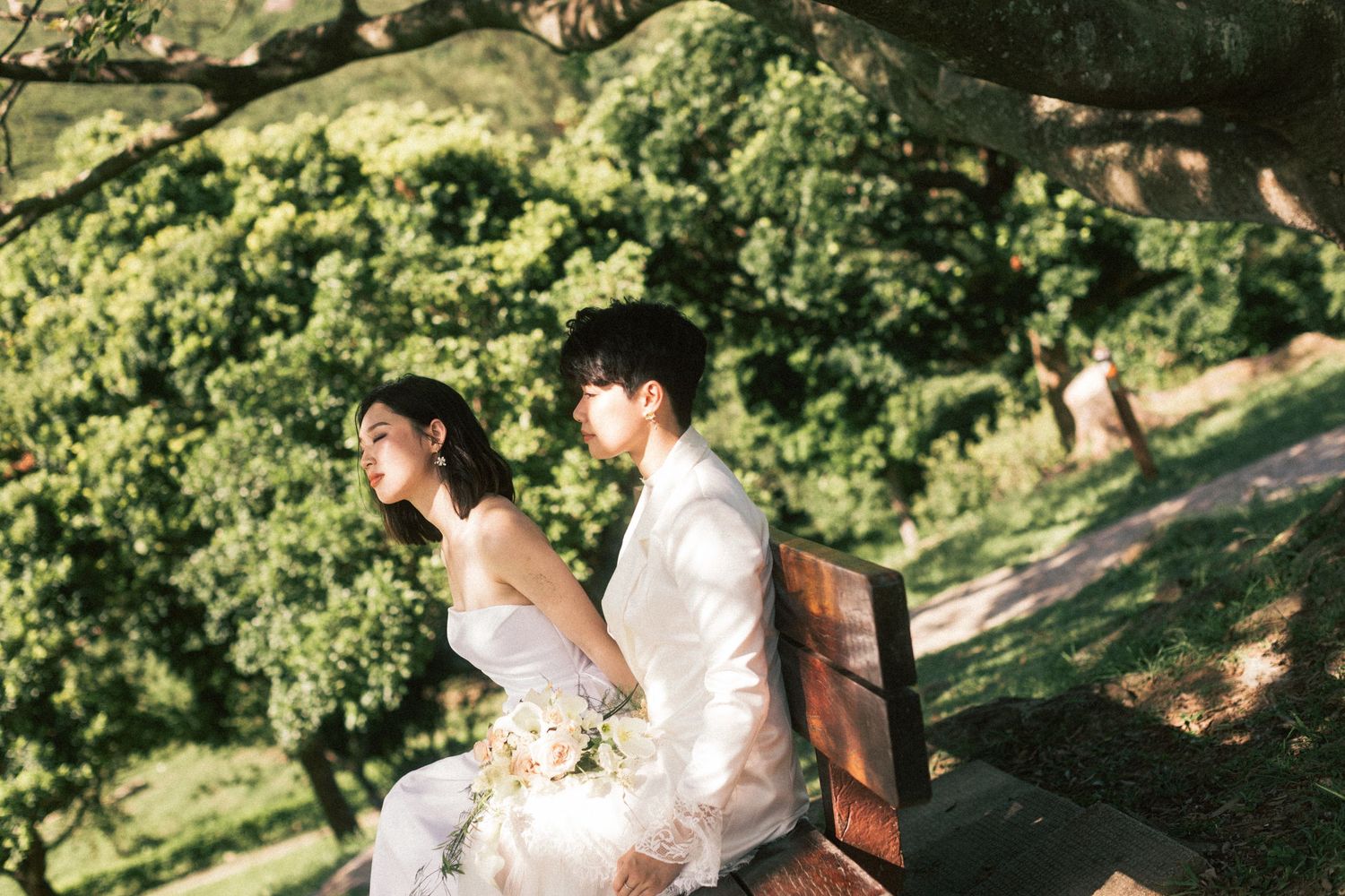 Two brides sit back-to-back on a wooden bench surrounded by lush green trees, one holding a white bouquet.