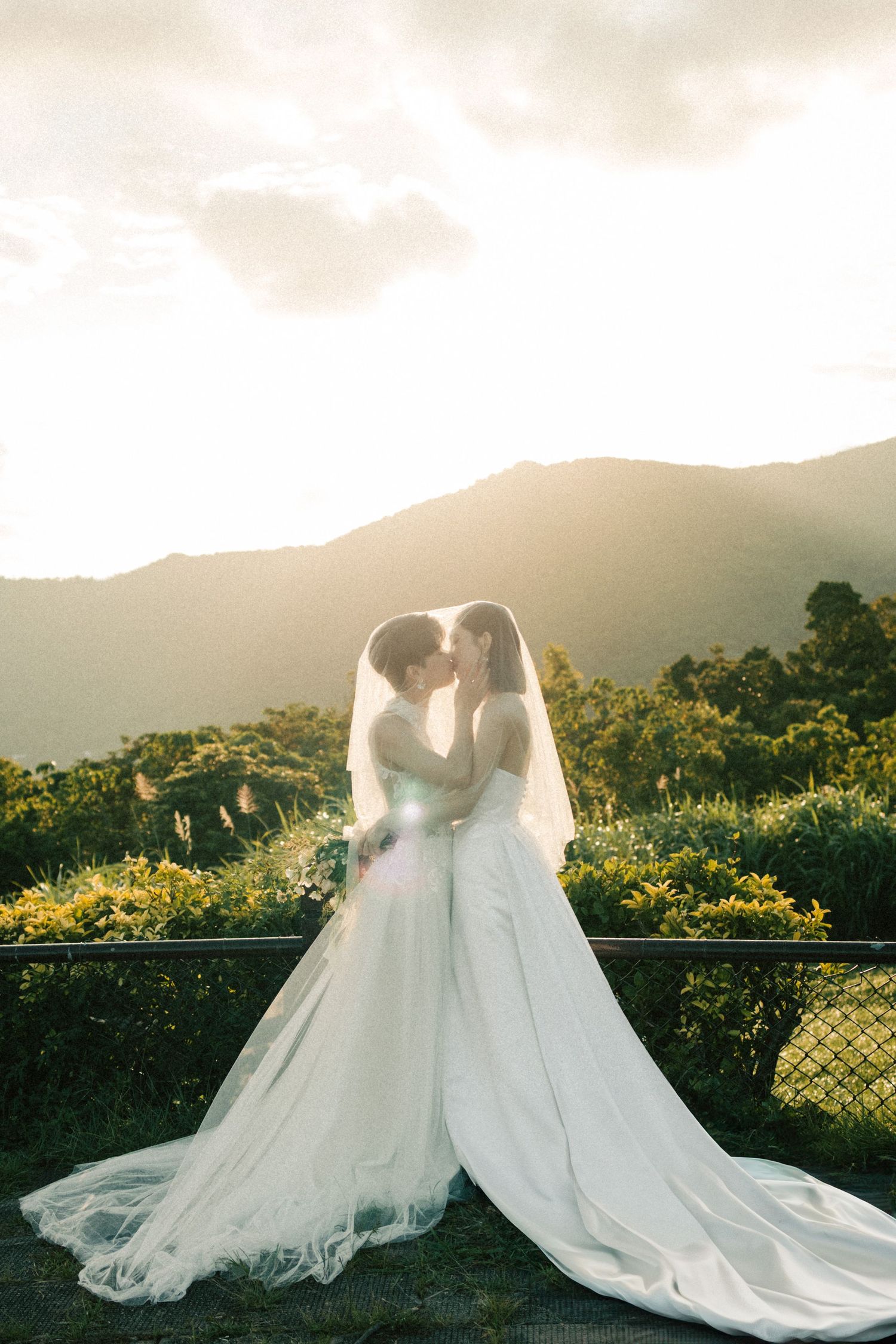 Two brides in flowing white gowns kiss under veils with a scenic mountain sunset.
