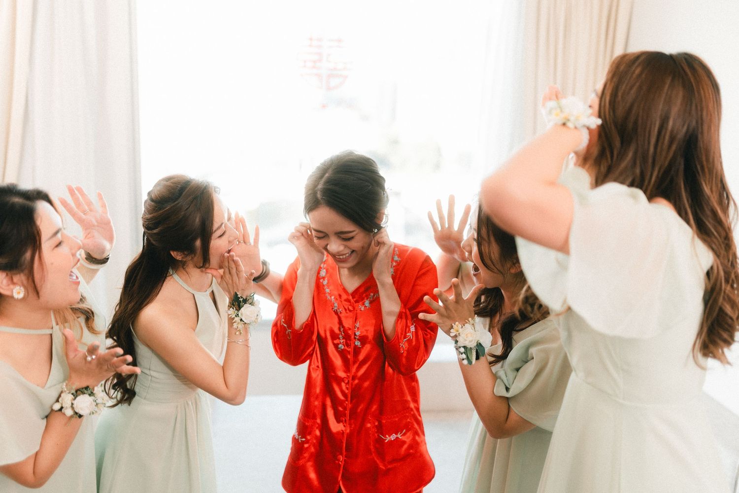 Emotional bride in red qipao laughs with bridesmaids in white dresses during wedding preparations.