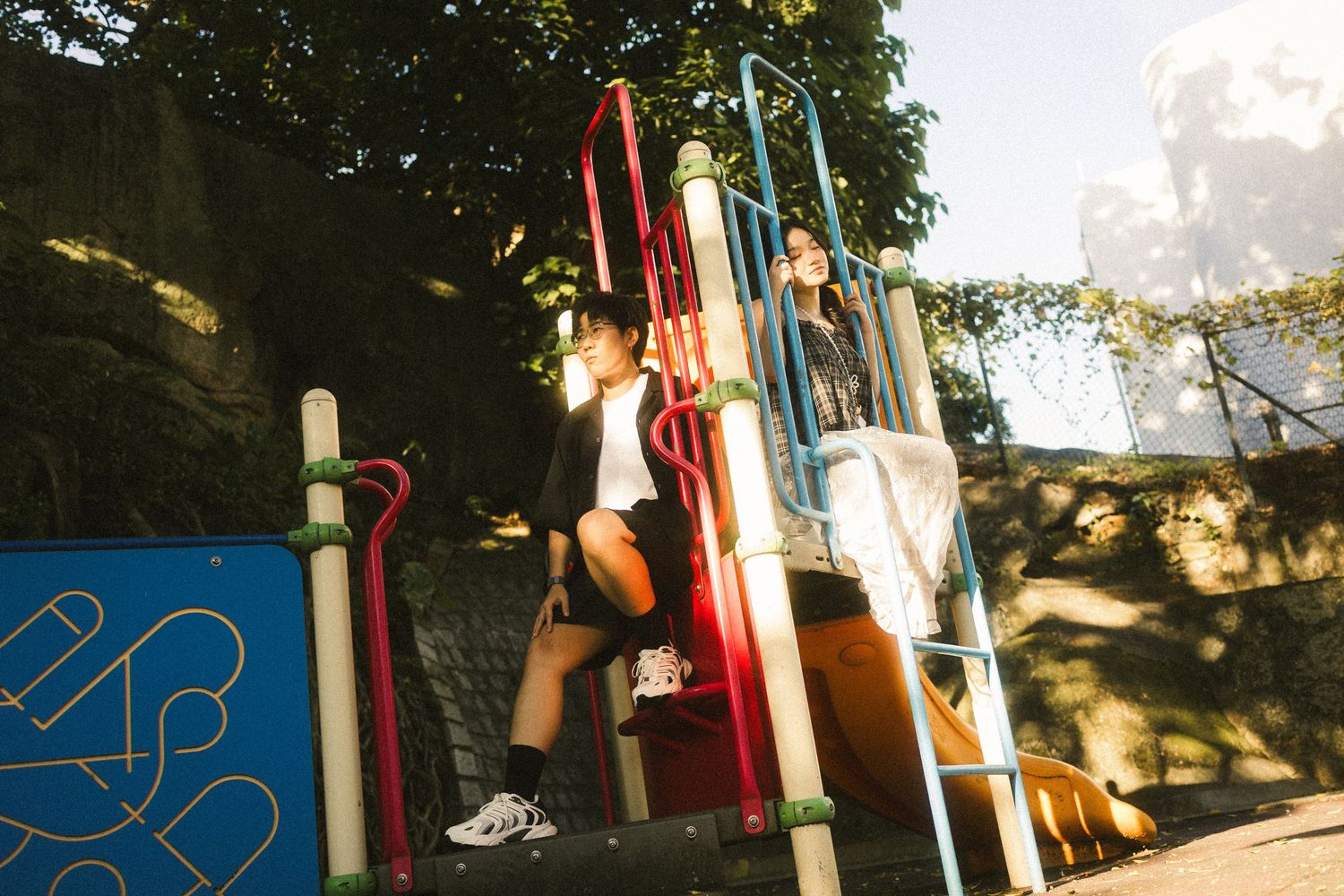 Two young people climbing colorful playground equipment in a sunny tree-shaded park.