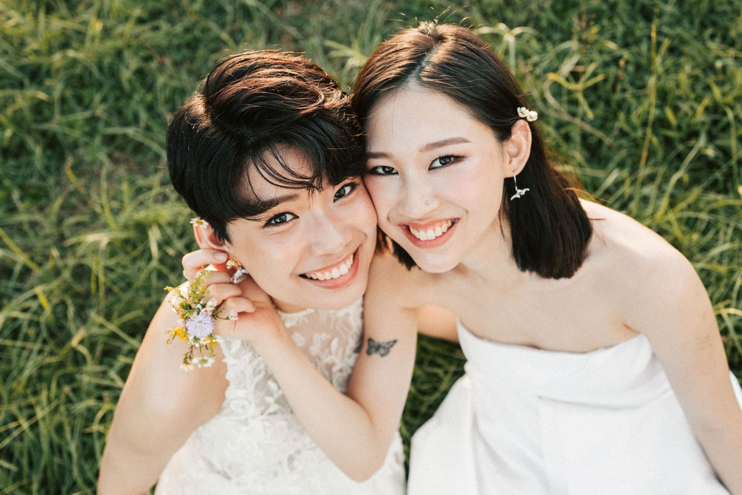 Two smiling brides in white wedding dresses pose together on a lush green grass background.