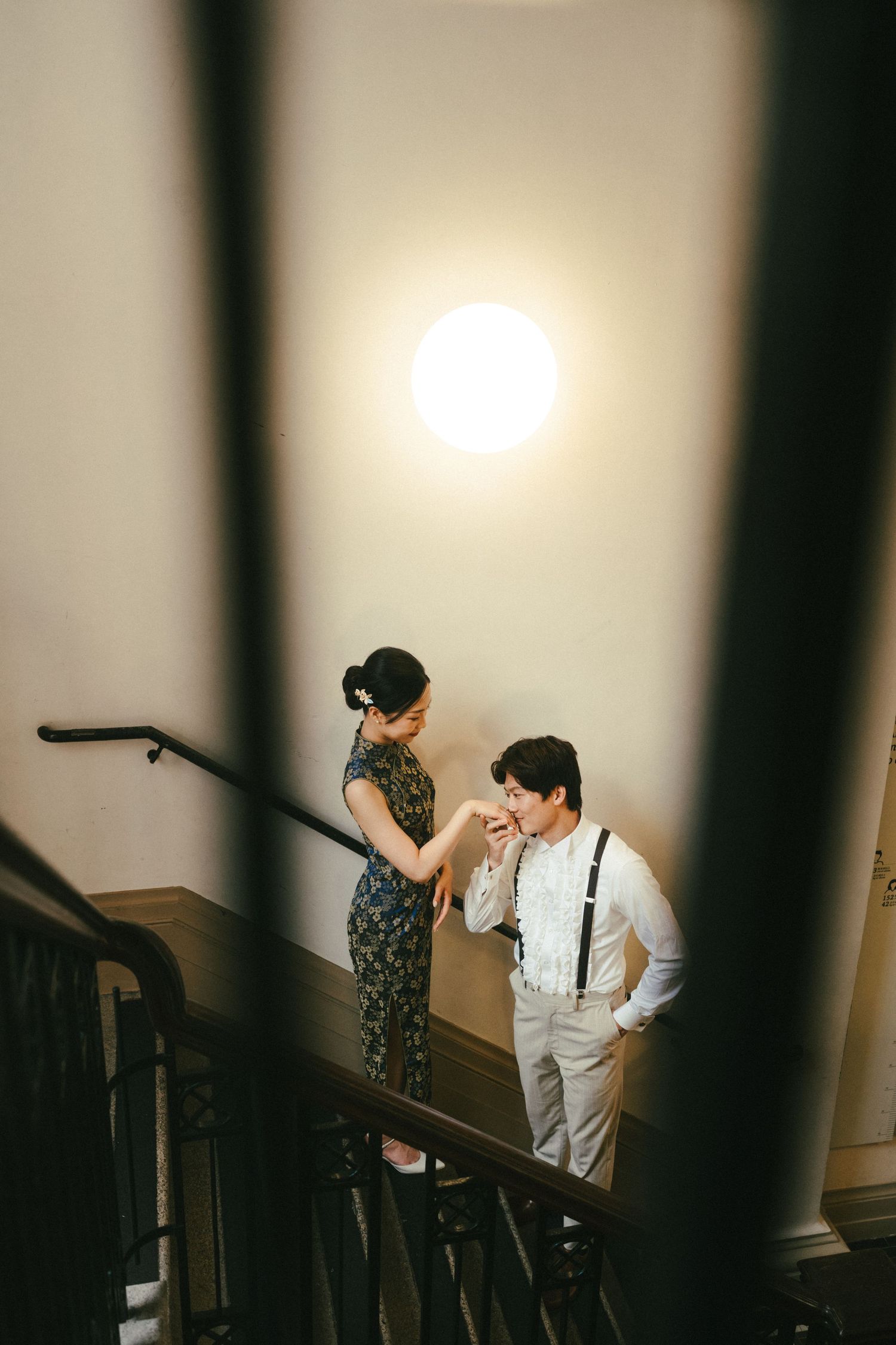 Couple holds hands on grand staircase beneath warm glowing light in elegant interior setting.