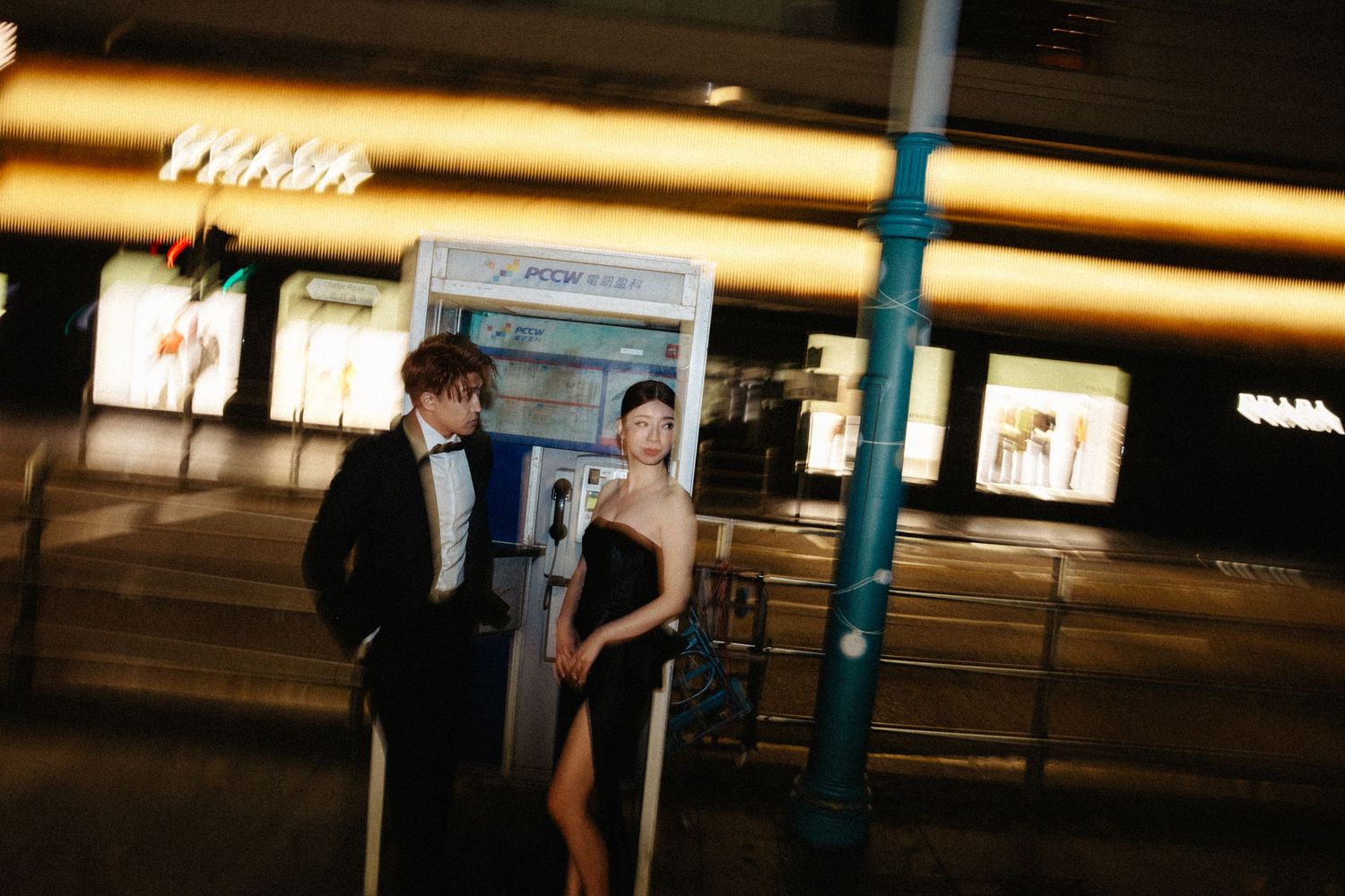 An elegant couple in formal attire stands by a phone booth on a city street at night.