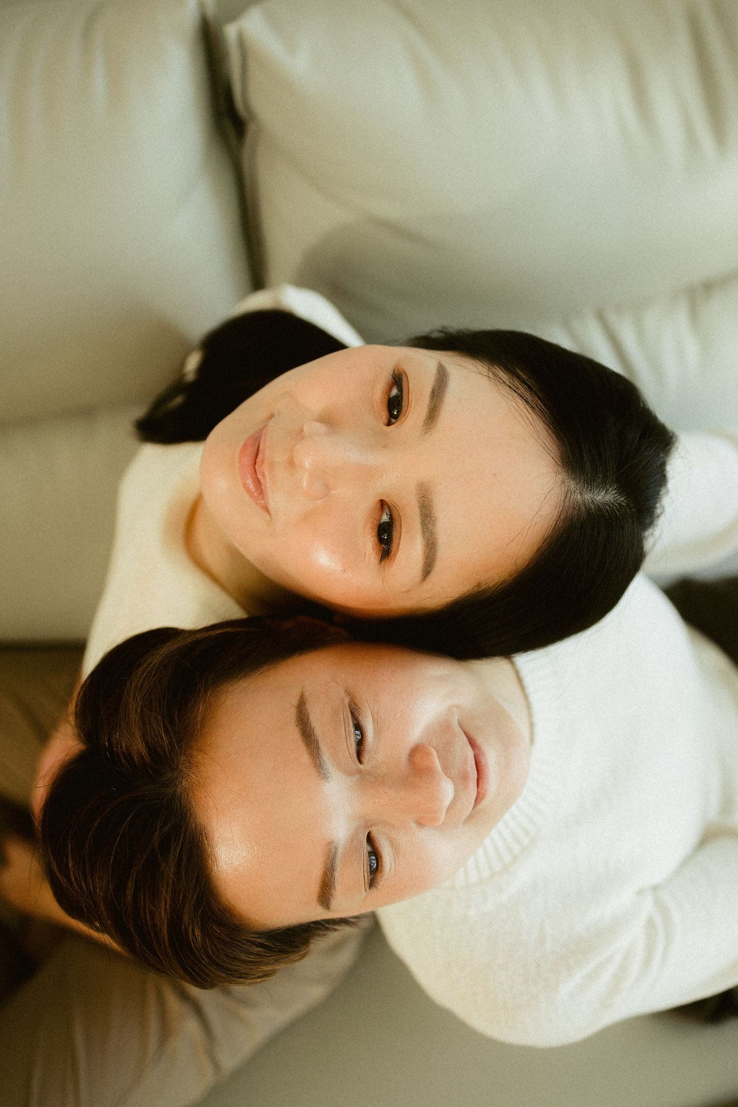 Two Asian women lean heads together smiling at camera from above on white leather sofa.