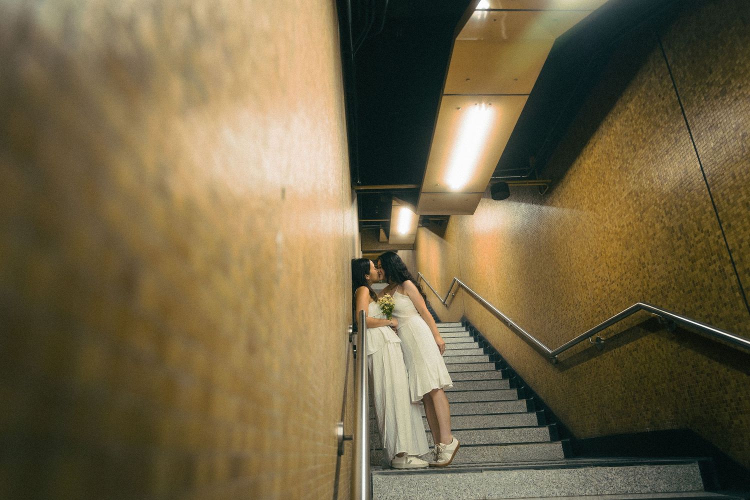 Woman in white dress stands on underground staircase between gold-toned walls under warm light.
