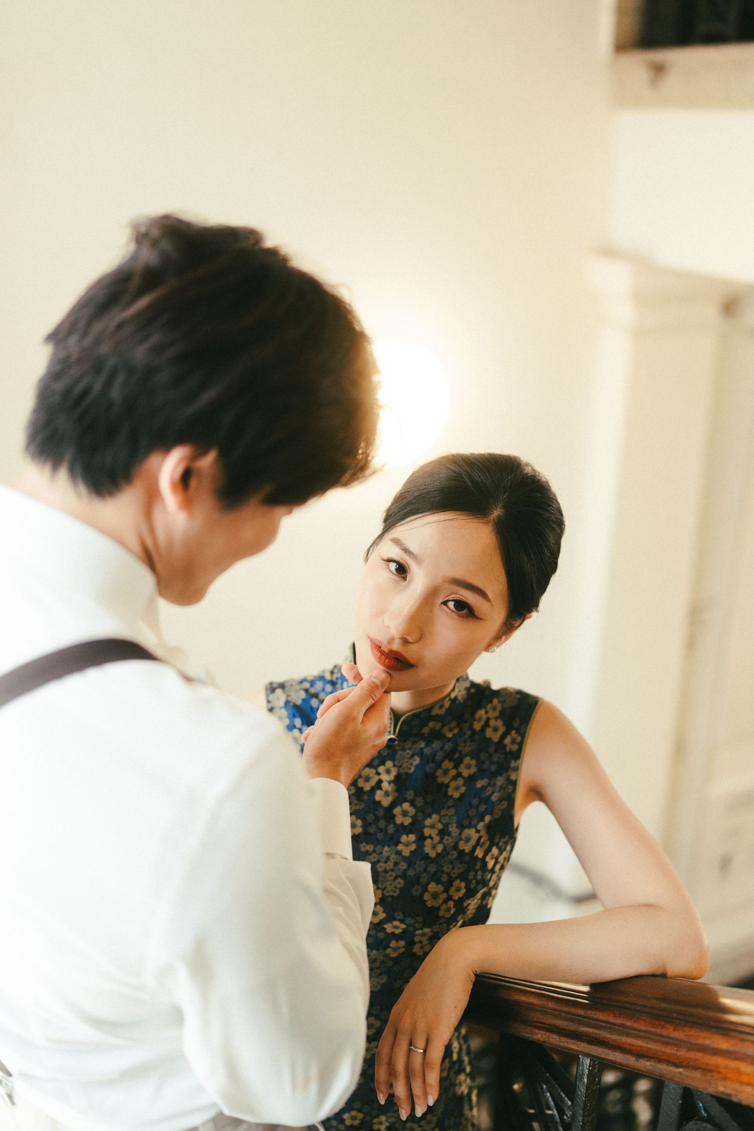 Groom tenderly touches brides lips as she leans on wooden banister in vintage interior.