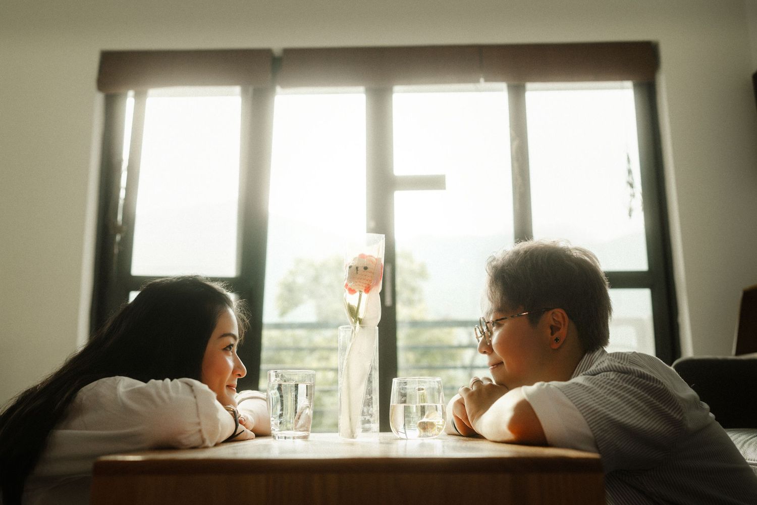 Two young people sitting across from each other at a wooden table near a bright window indoors.