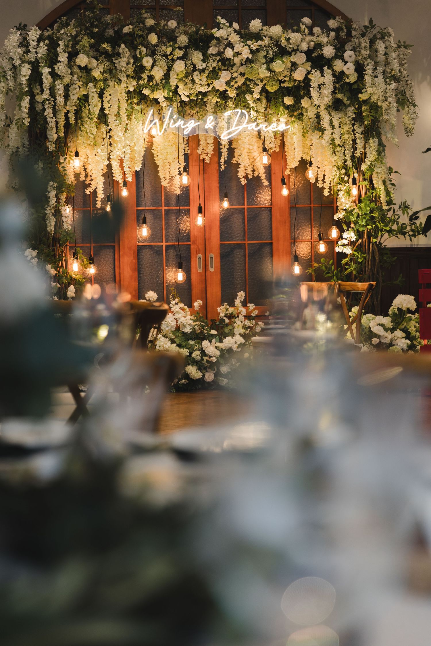 Dreamy wedding reception backdrop with neon sign, cascading white flowers, and warm Edison lights.