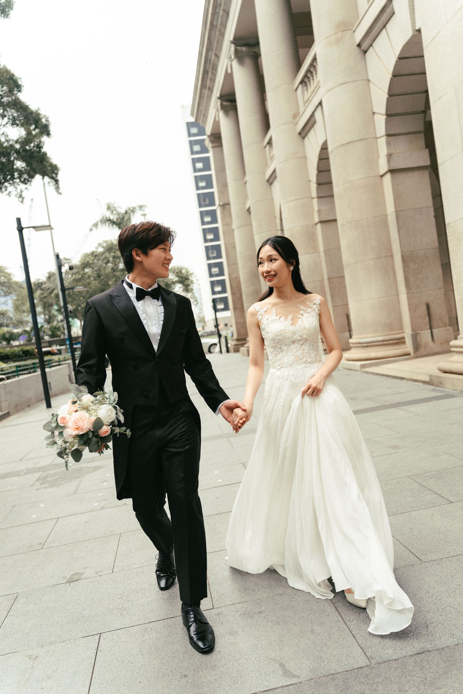 An elegant Asian couple in wedding attire walk hand-in-hand past grand stone columns, smiling at each other.