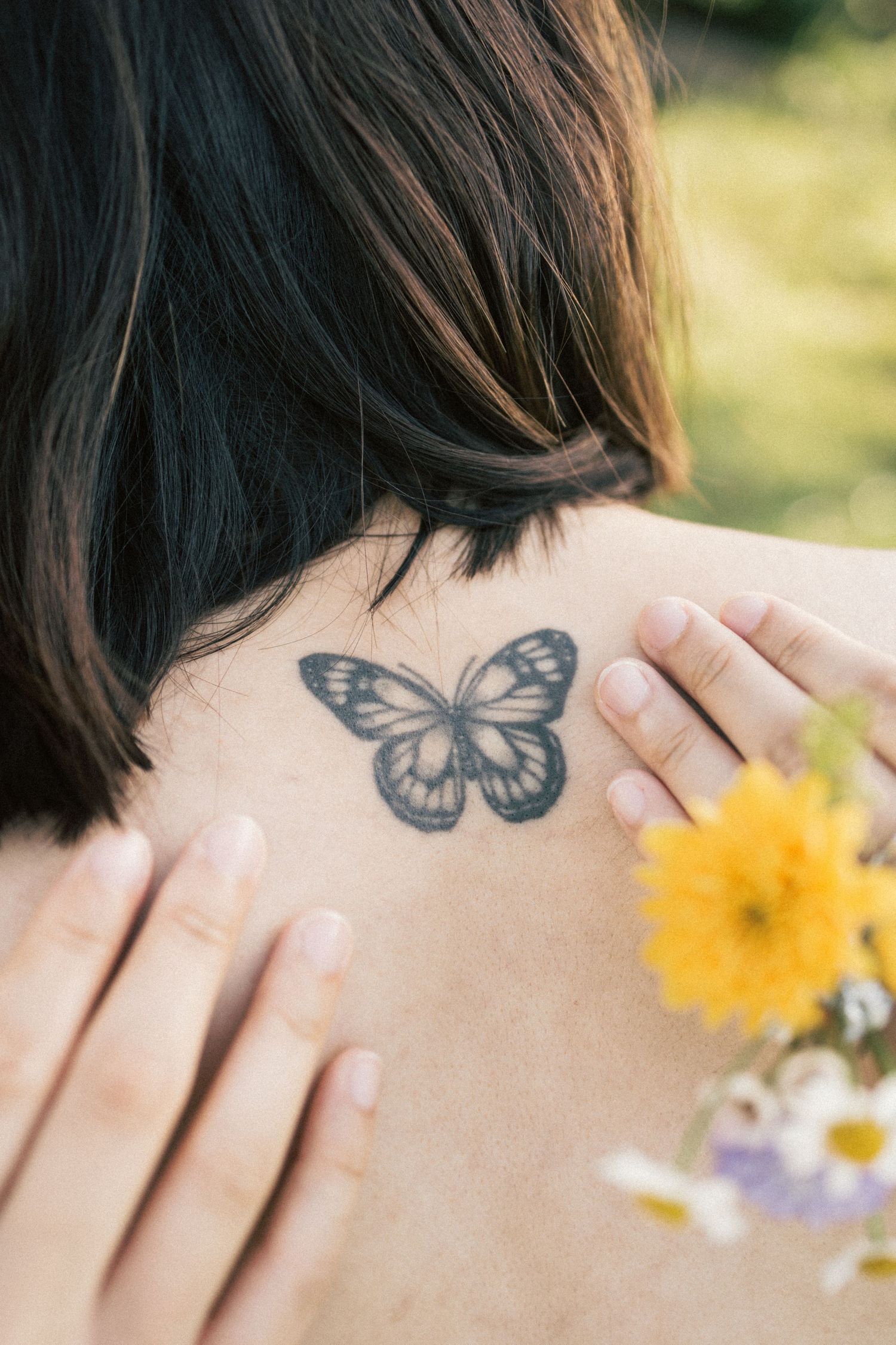 Close-up of a butterfly tattoo on a woman's upper back with yellow flowers nearby.