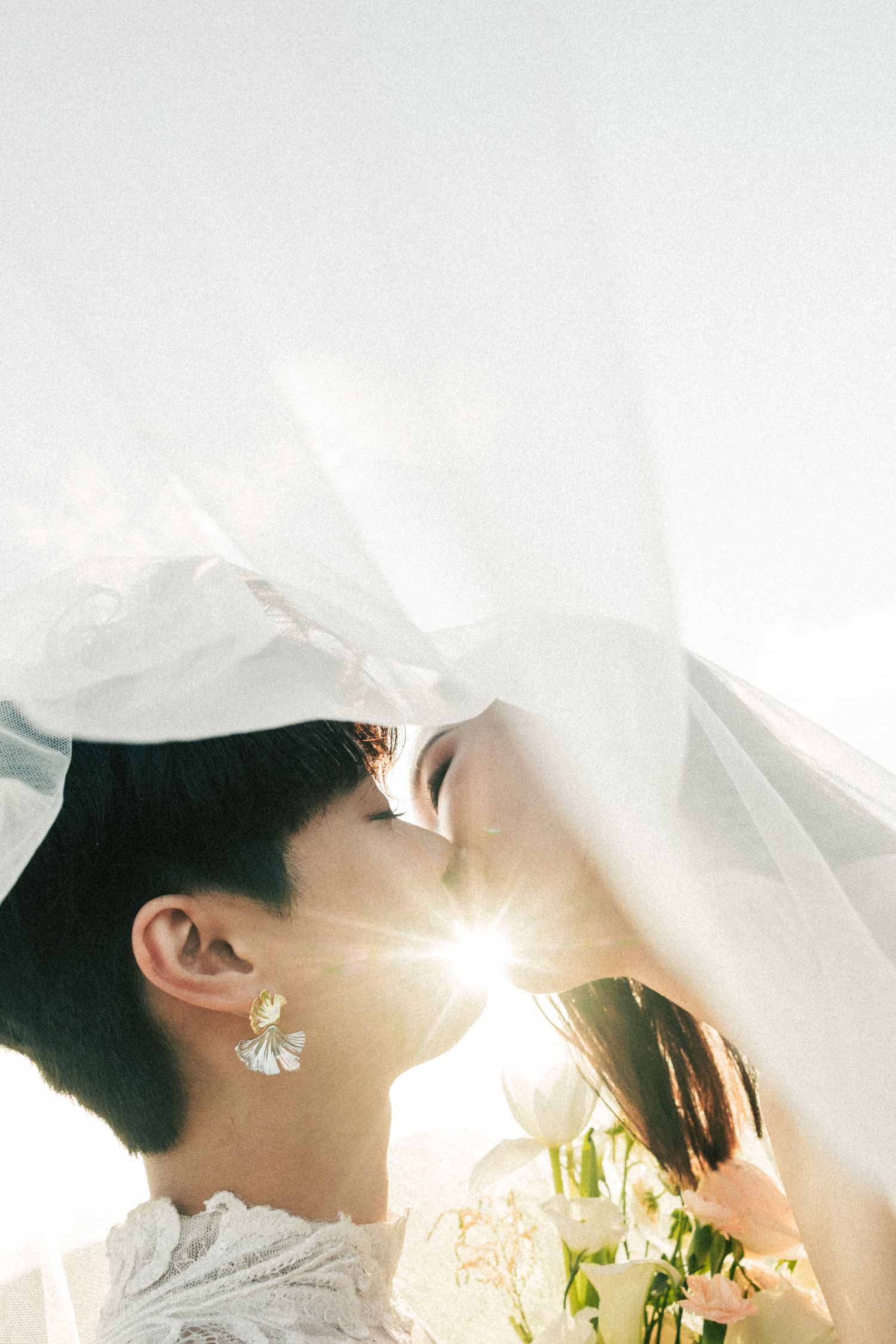 A bride with short hair holds flowers under a flowing veil with sunlight streaming through.