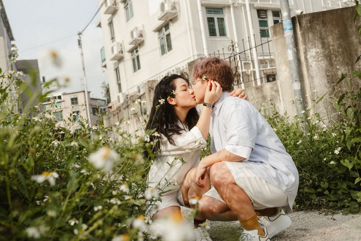 Couple sharing a kiss while crouching among white wildflowers in an outdoor urban garden setting.