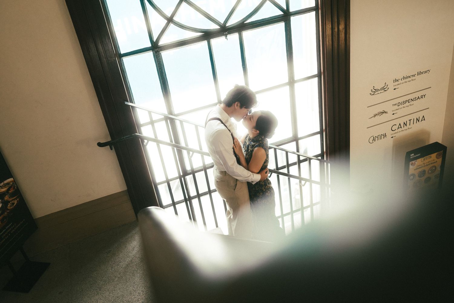 Couple shares romantic kiss silhouetted against large arched window with bright natural light.