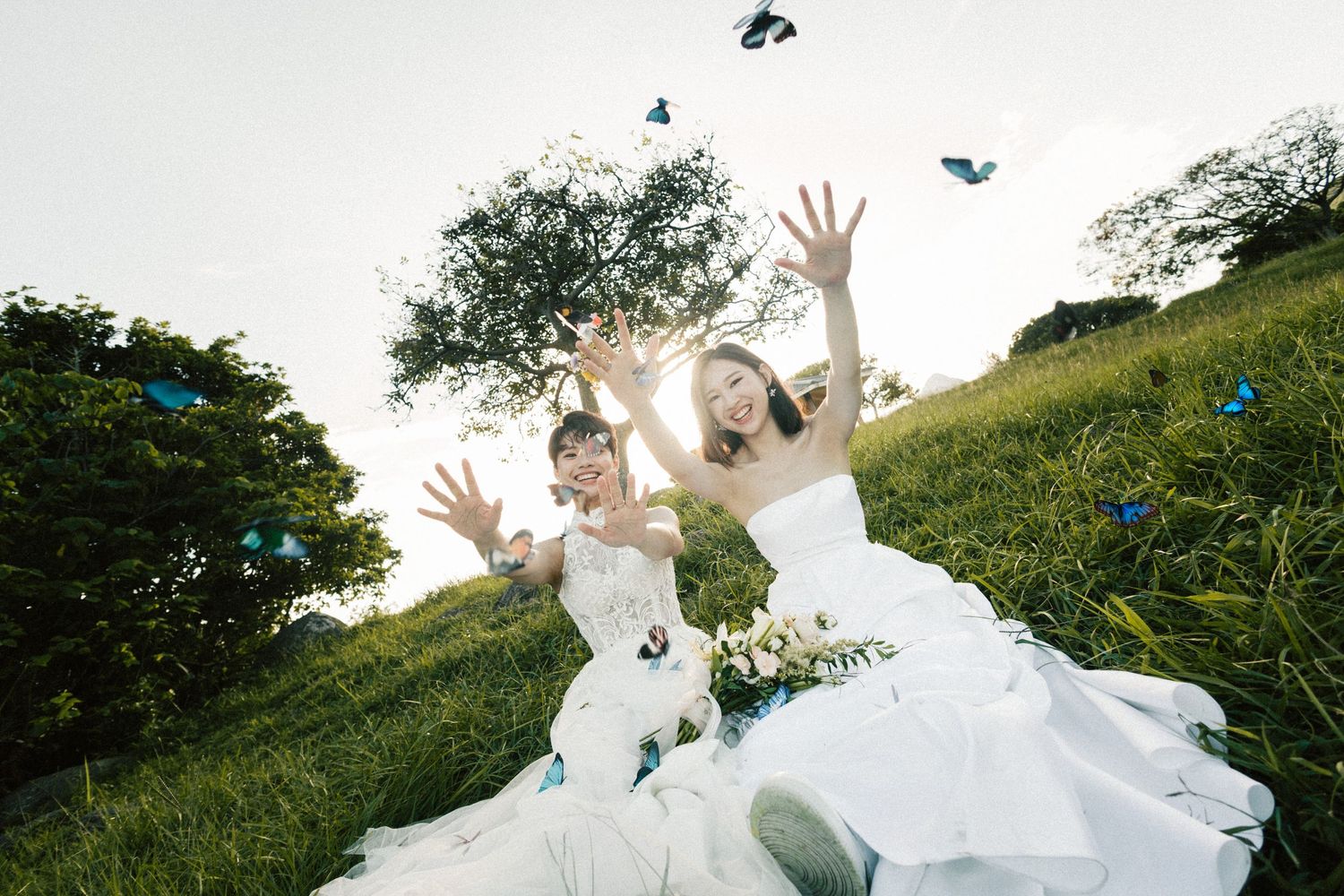 Two joyful brides in white gowns toss blue butterflies into the air on a hillside.
