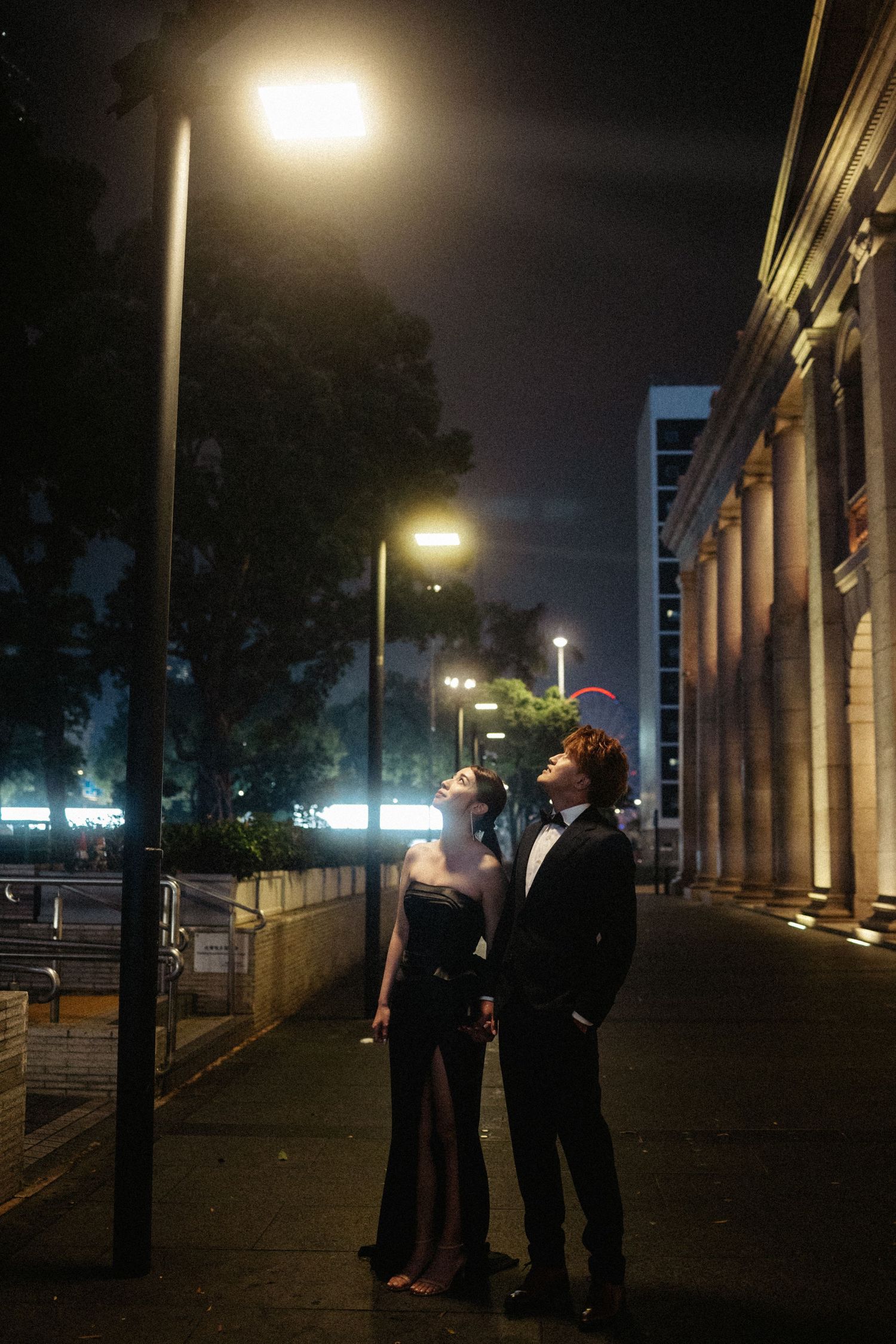 An elegant couple in formal attire gazes upward under a streetlight on a quiet night.