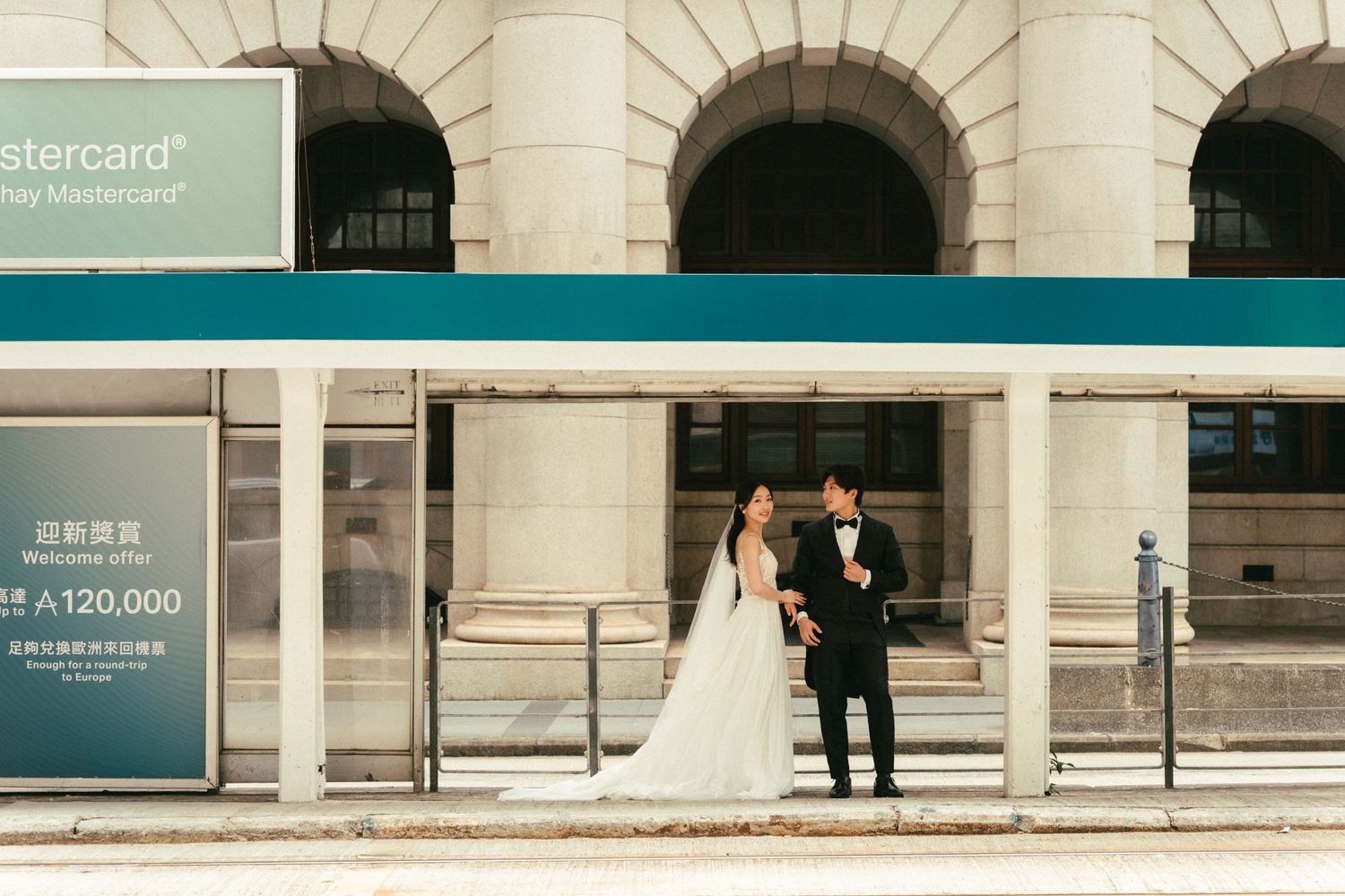 A bride and groom in formal wedding attire stand together at a city bus stop with urban architecture behind them.