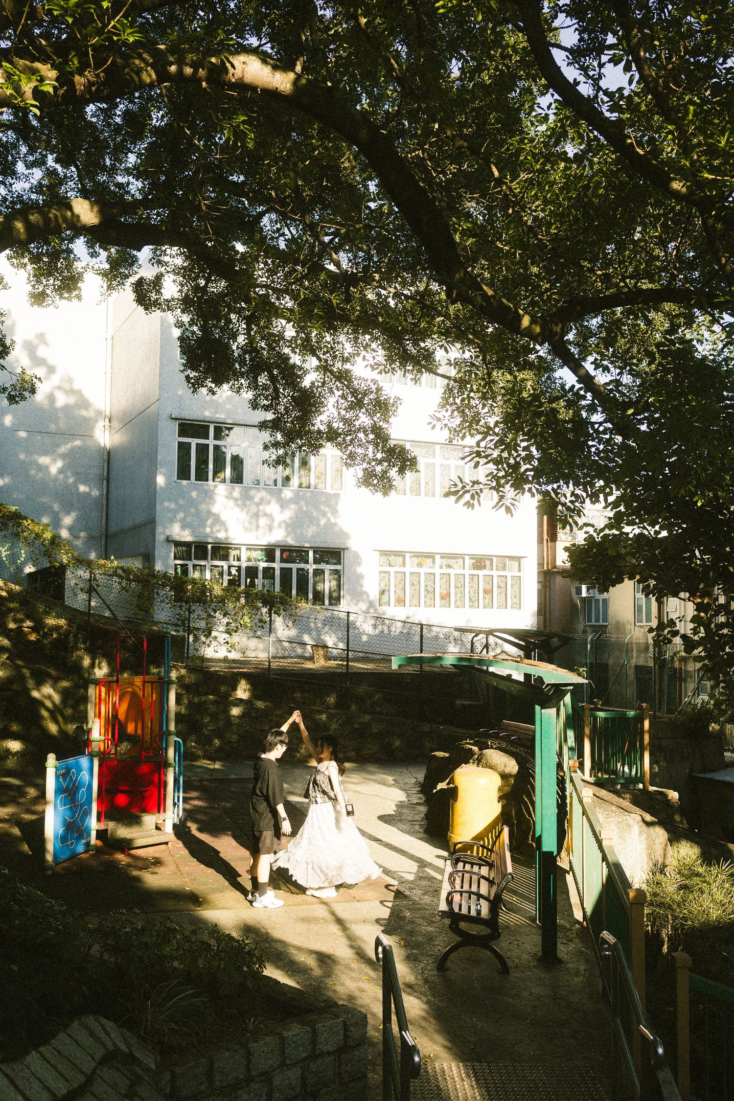 Wedding couple walking through a sunlit courtyard with lush trees and a white building backdrop.