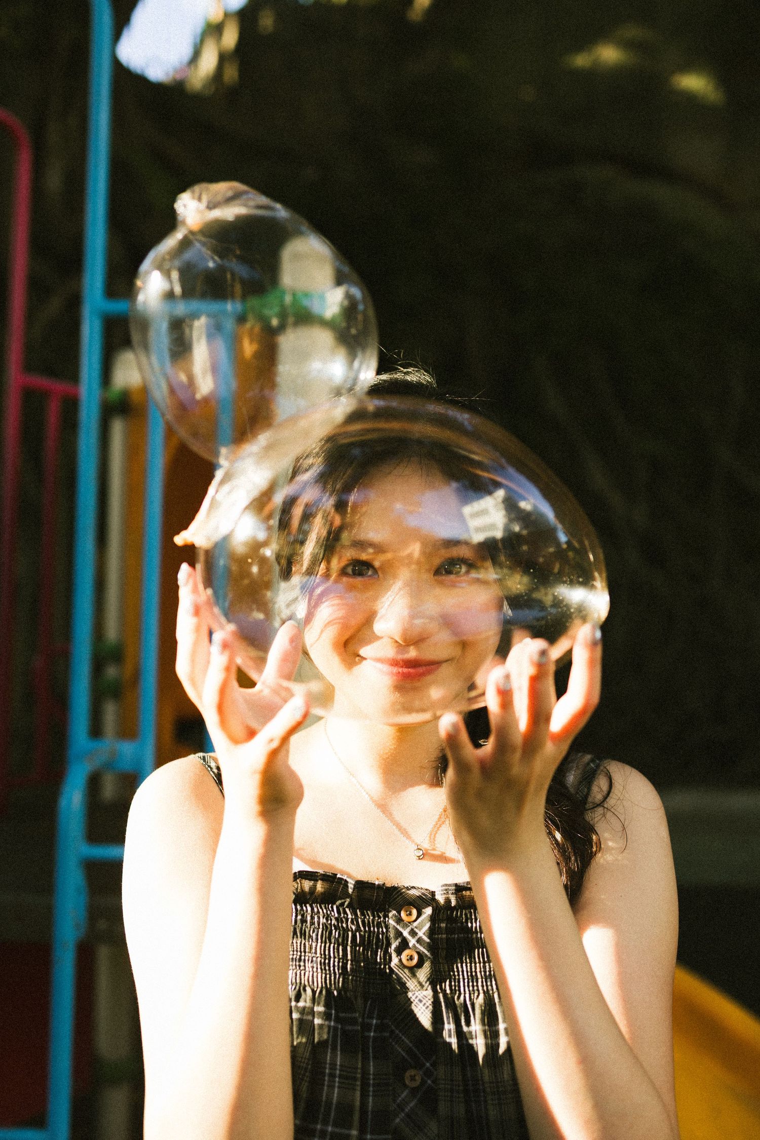 Smiling young woman holding a large transparent bubble in front of her face at a sunny playground.