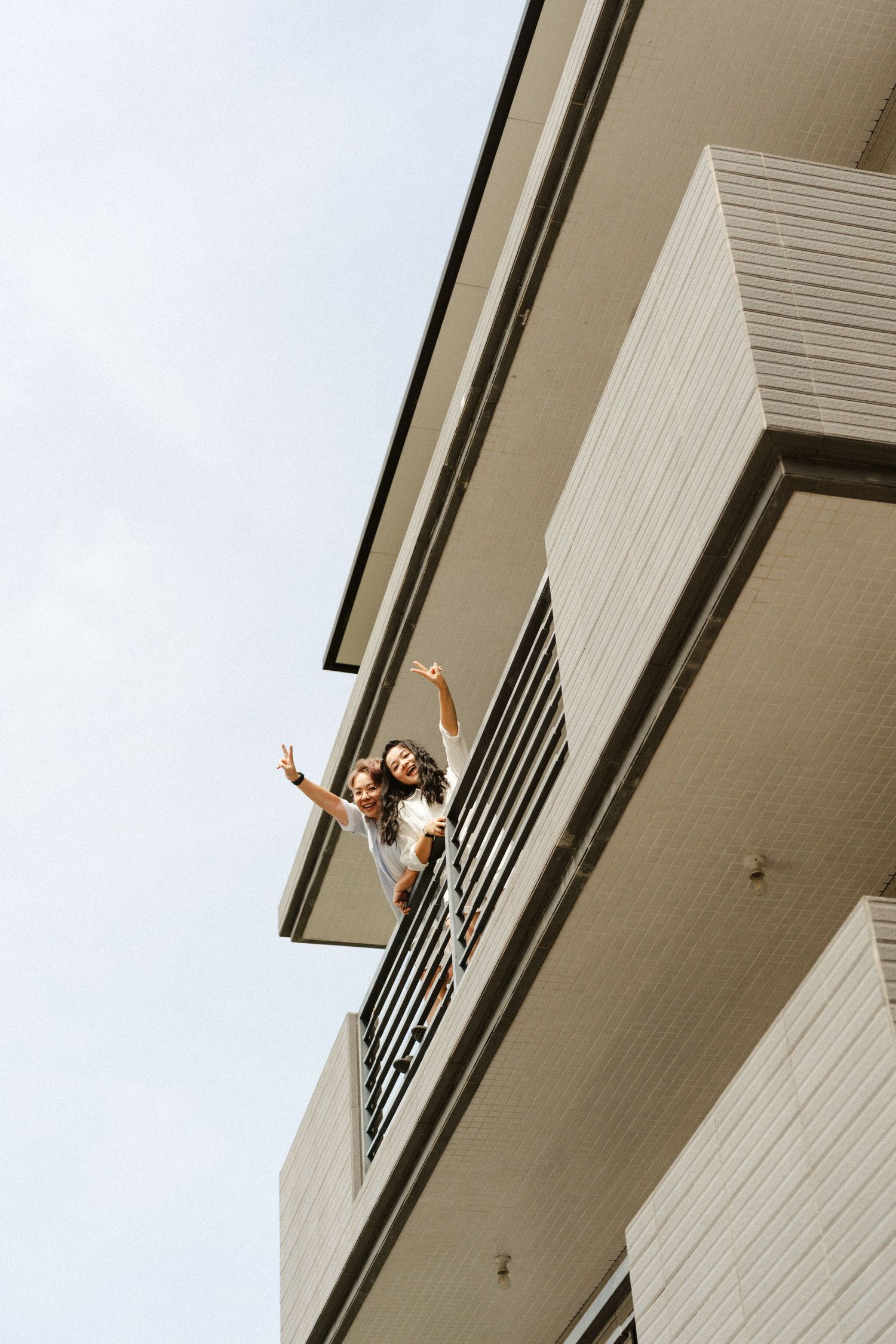 Two people waving from a high-rise apartment balcony against a clear blue sky.