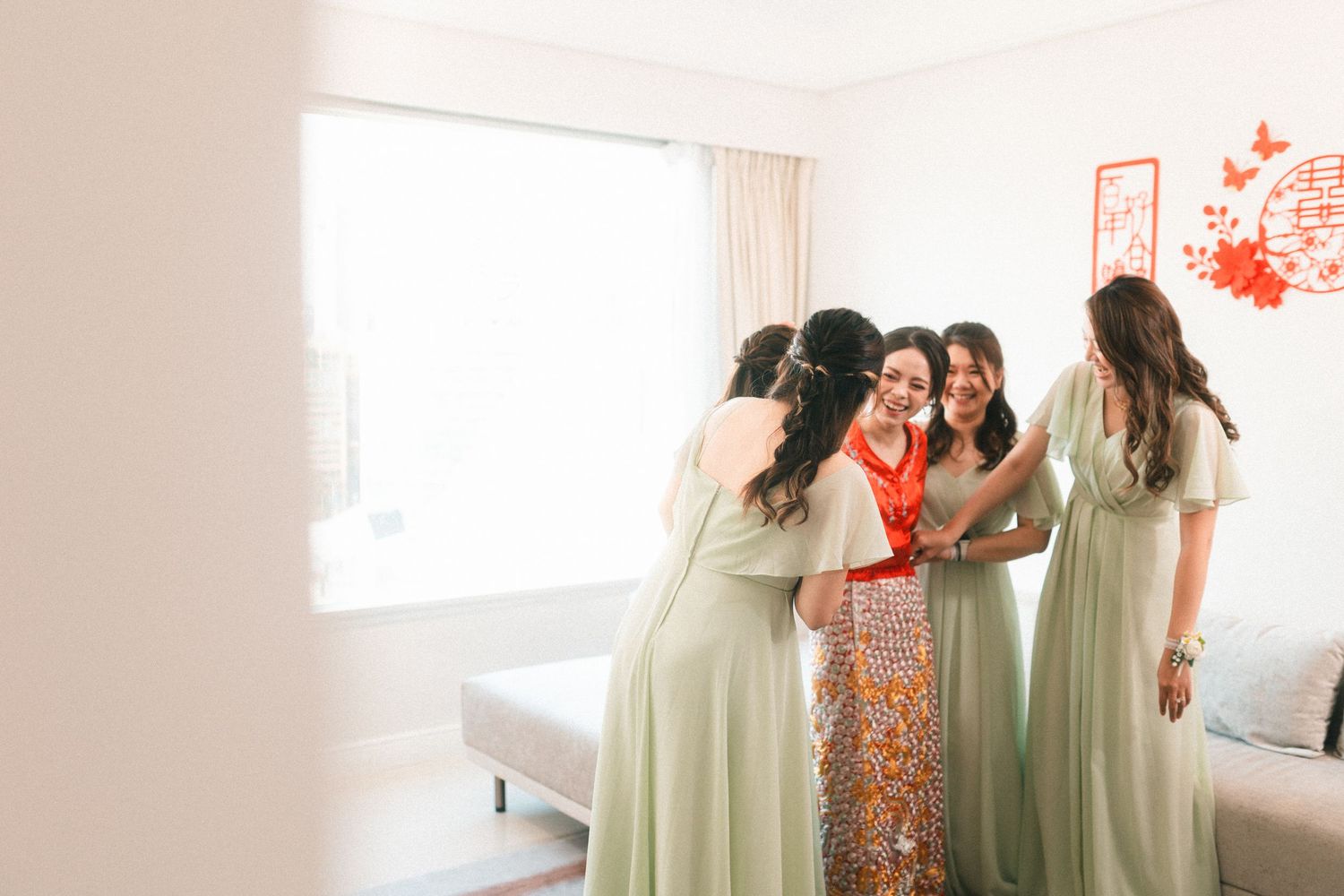 Bridesmaids in sage green dresses share a warm group hug with the bride during wedding preparations in a bright room.