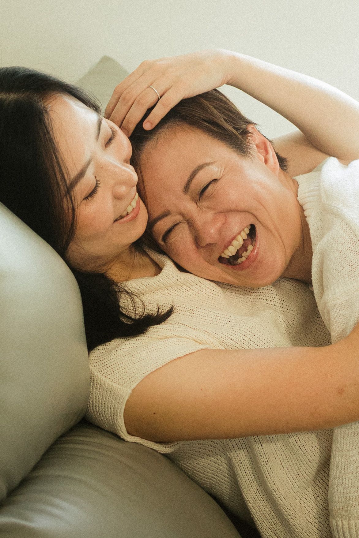 Two Asian women laughing joyfully while embracing on a white leather sofa together.