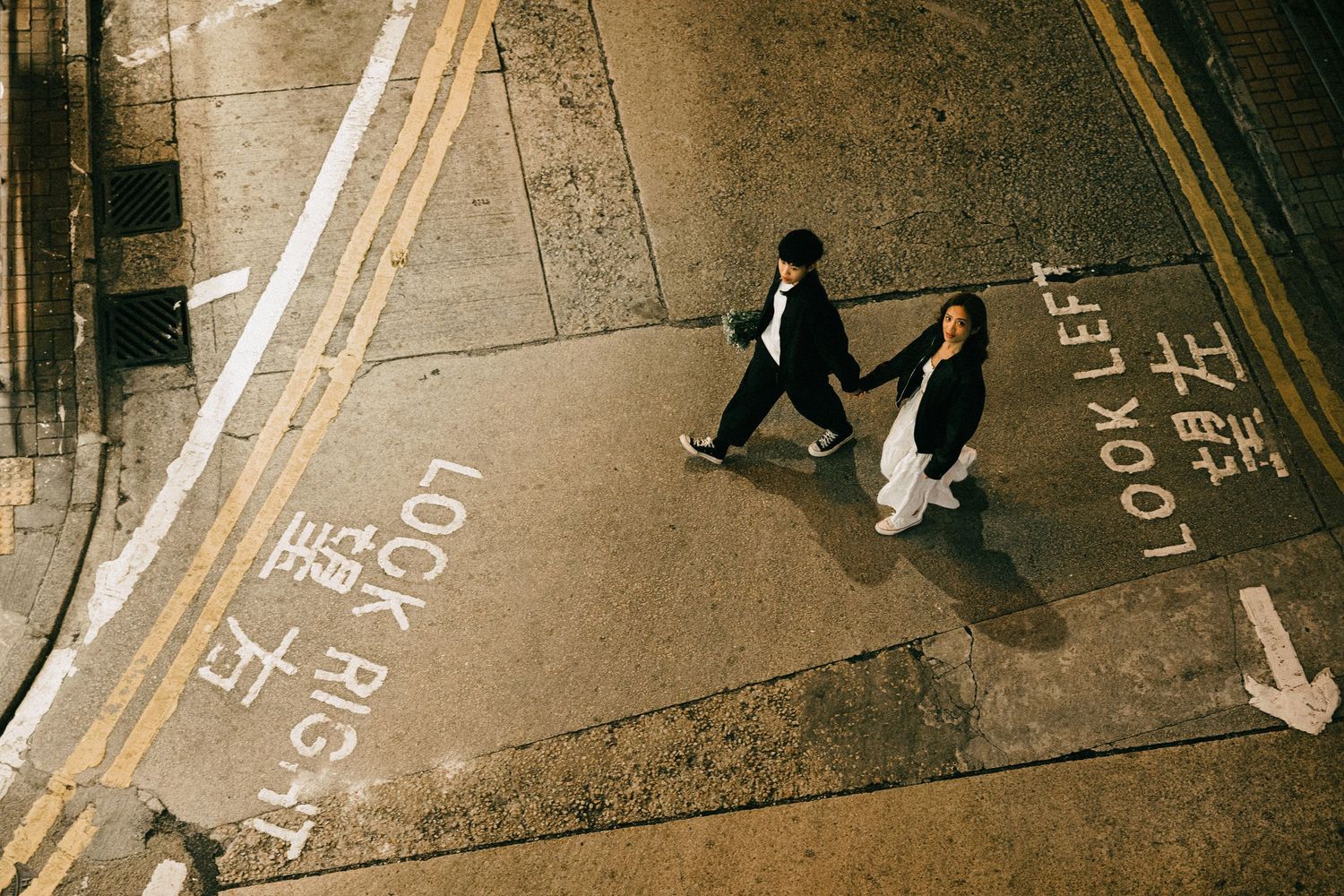 An aerial view of an Asian couple holding hands while crossing a city street crosswalk.