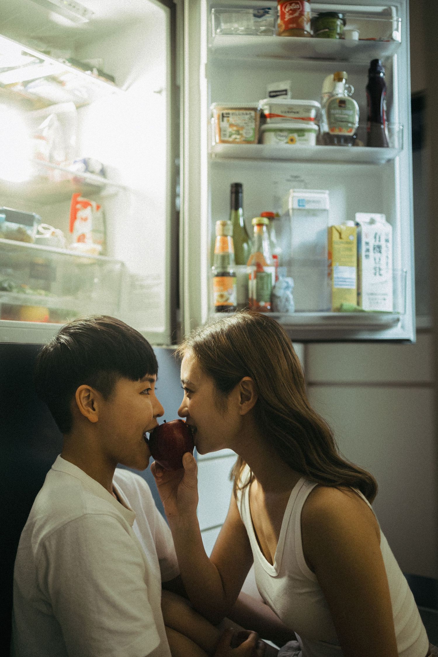 Couple sharing an apple in front of an open refrigerator at night.