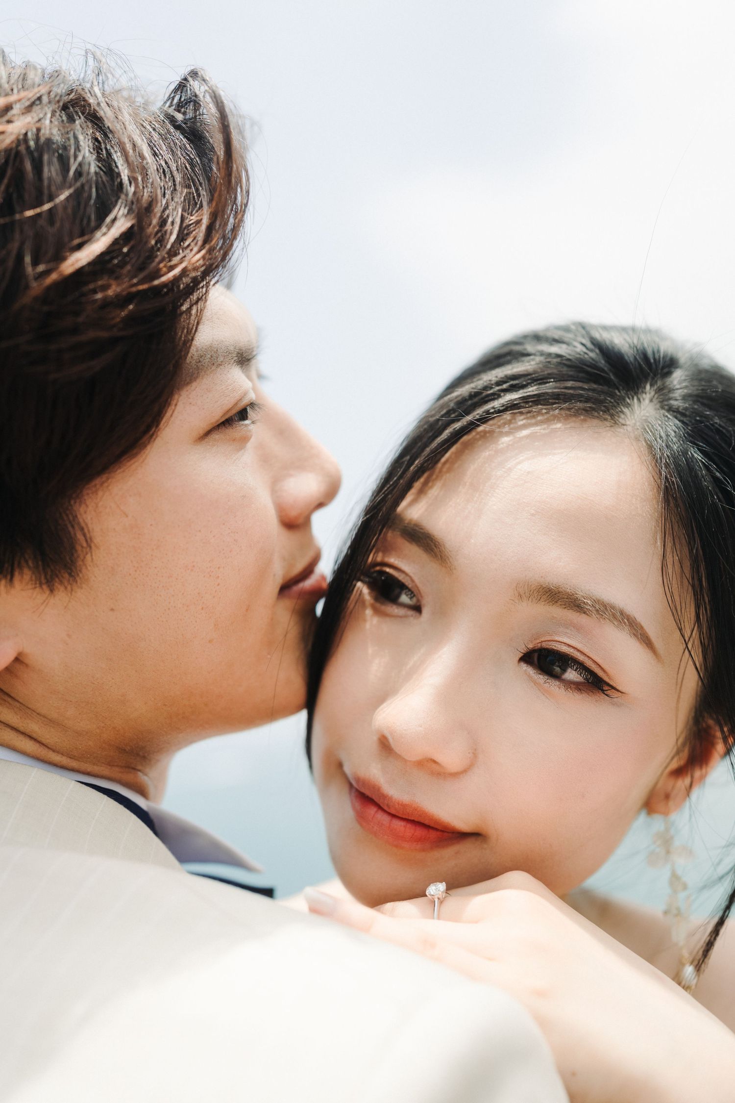 Groom gently kisses cheek of smiling Asian bride wearing engagement ring against soft sky background.