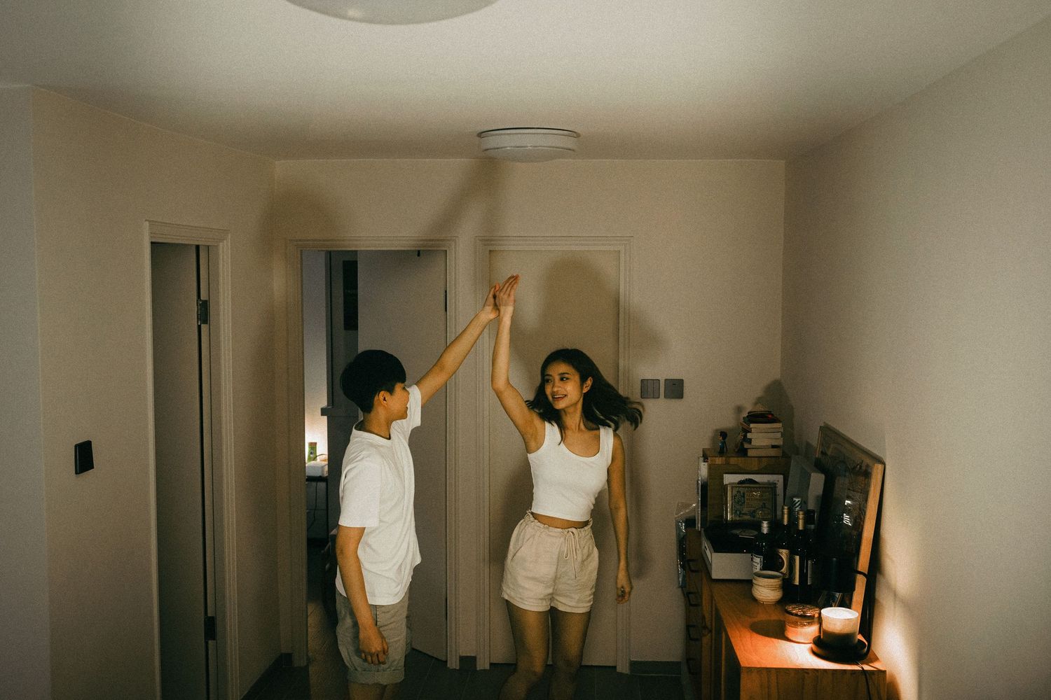 A young Asian couple playfully reaching up to touch the ceiling light in their cozy apartment.