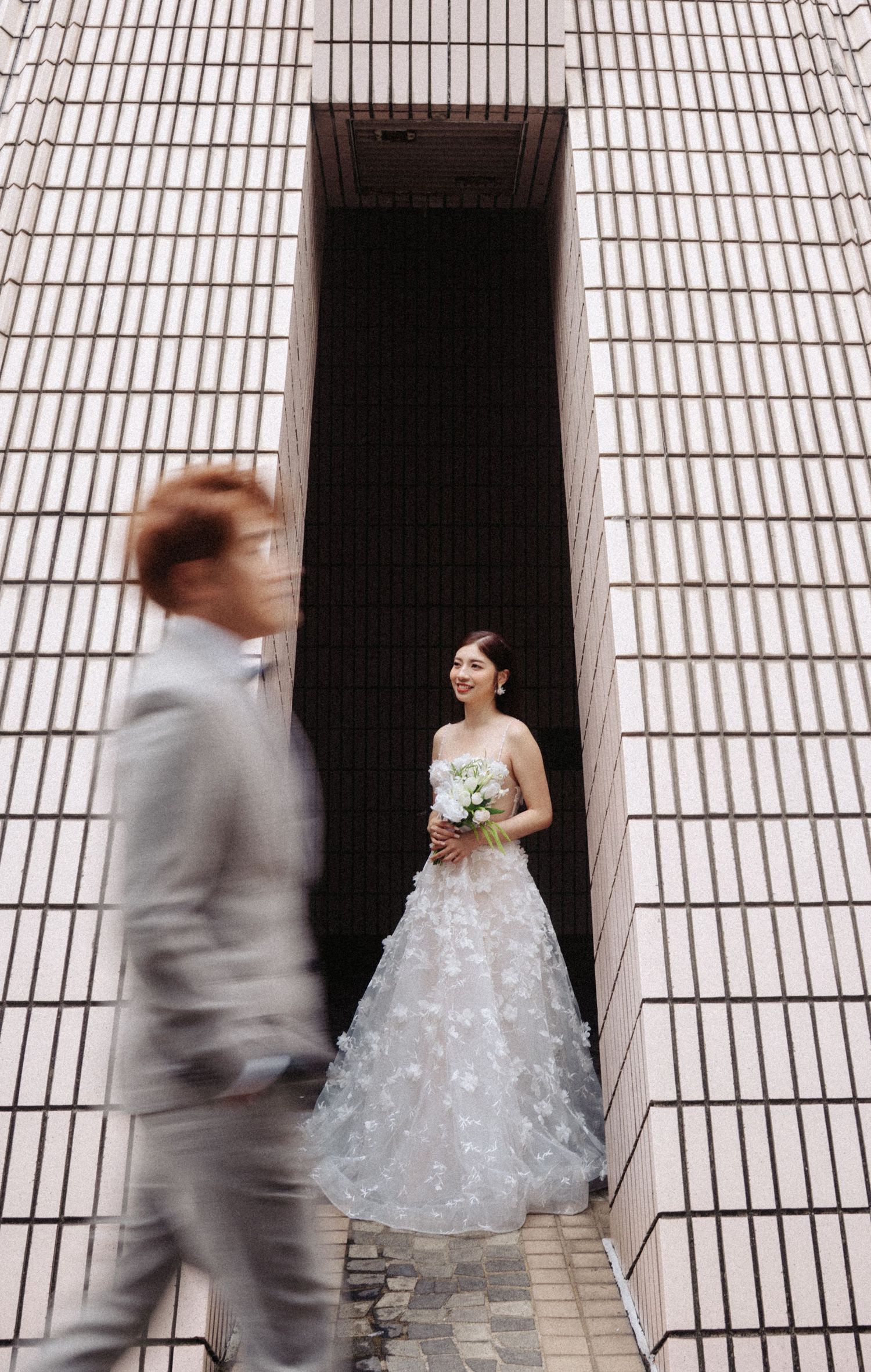 Bride in floral gown stands in narrow building gap as groom walks past in motion blur.