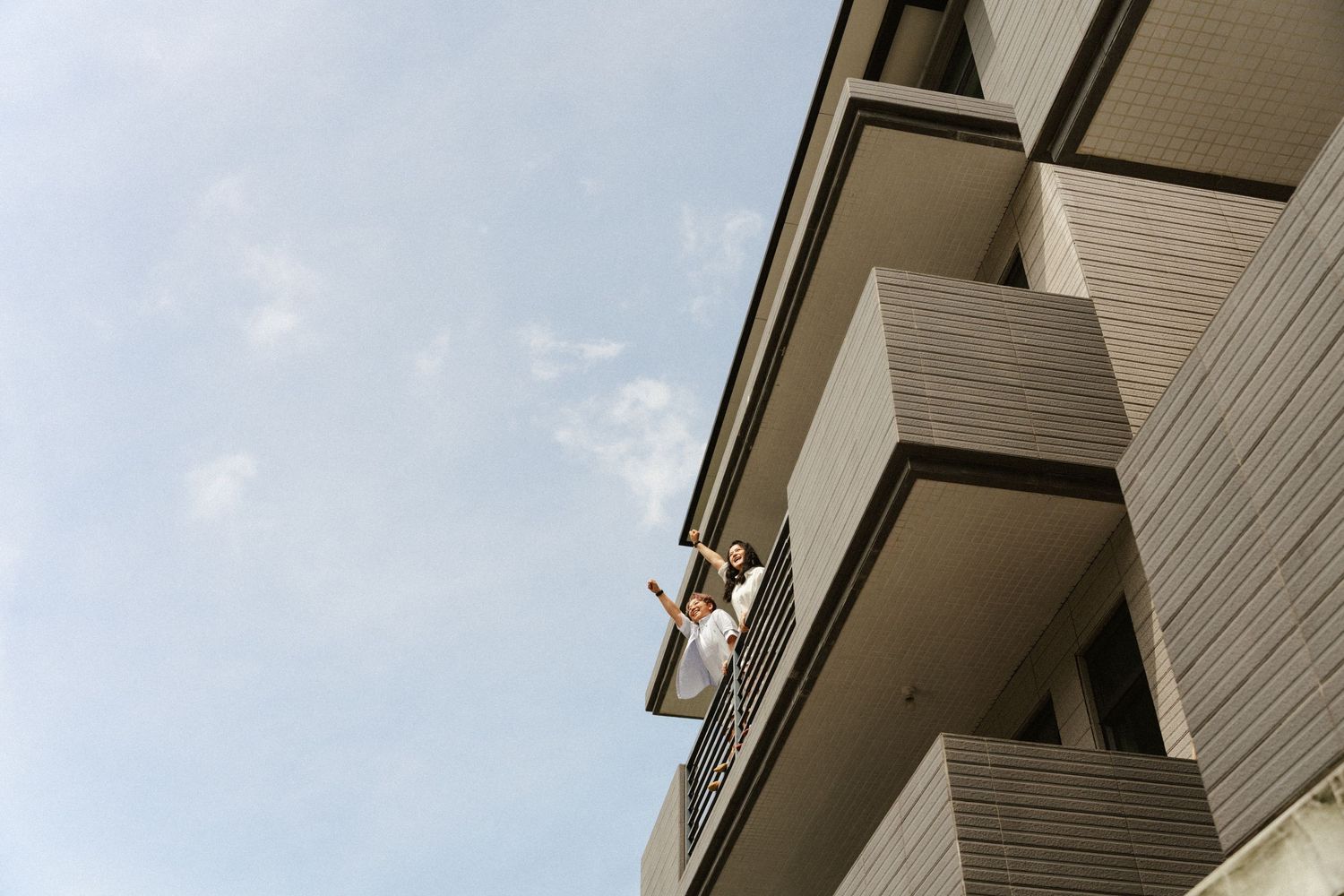 Person leaning over apartment balcony railing with arms raised against a bright blue sky.