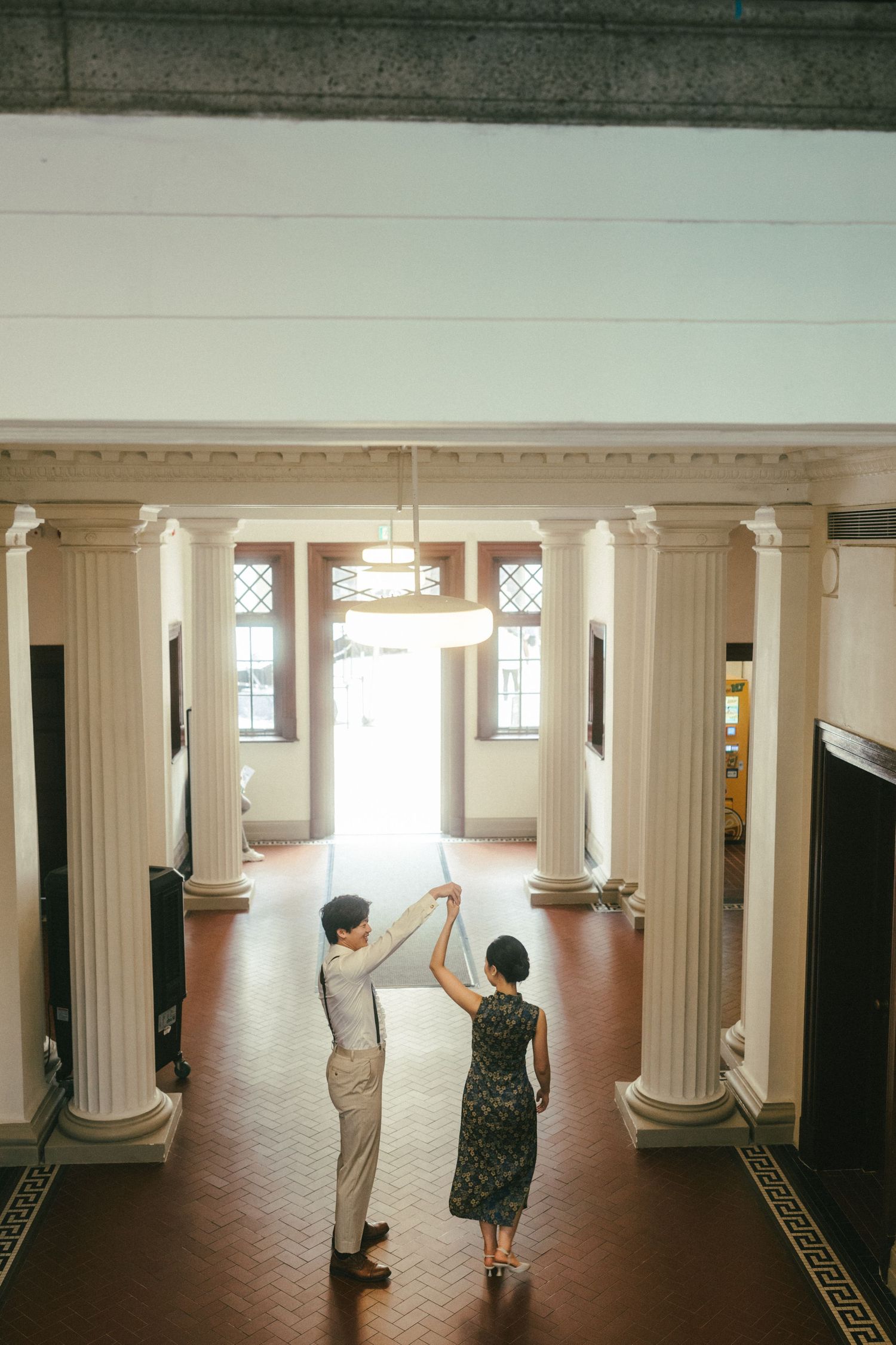 Couple dances playfully beneath grand chandelier surrounded by classical white columns in elegant hall.