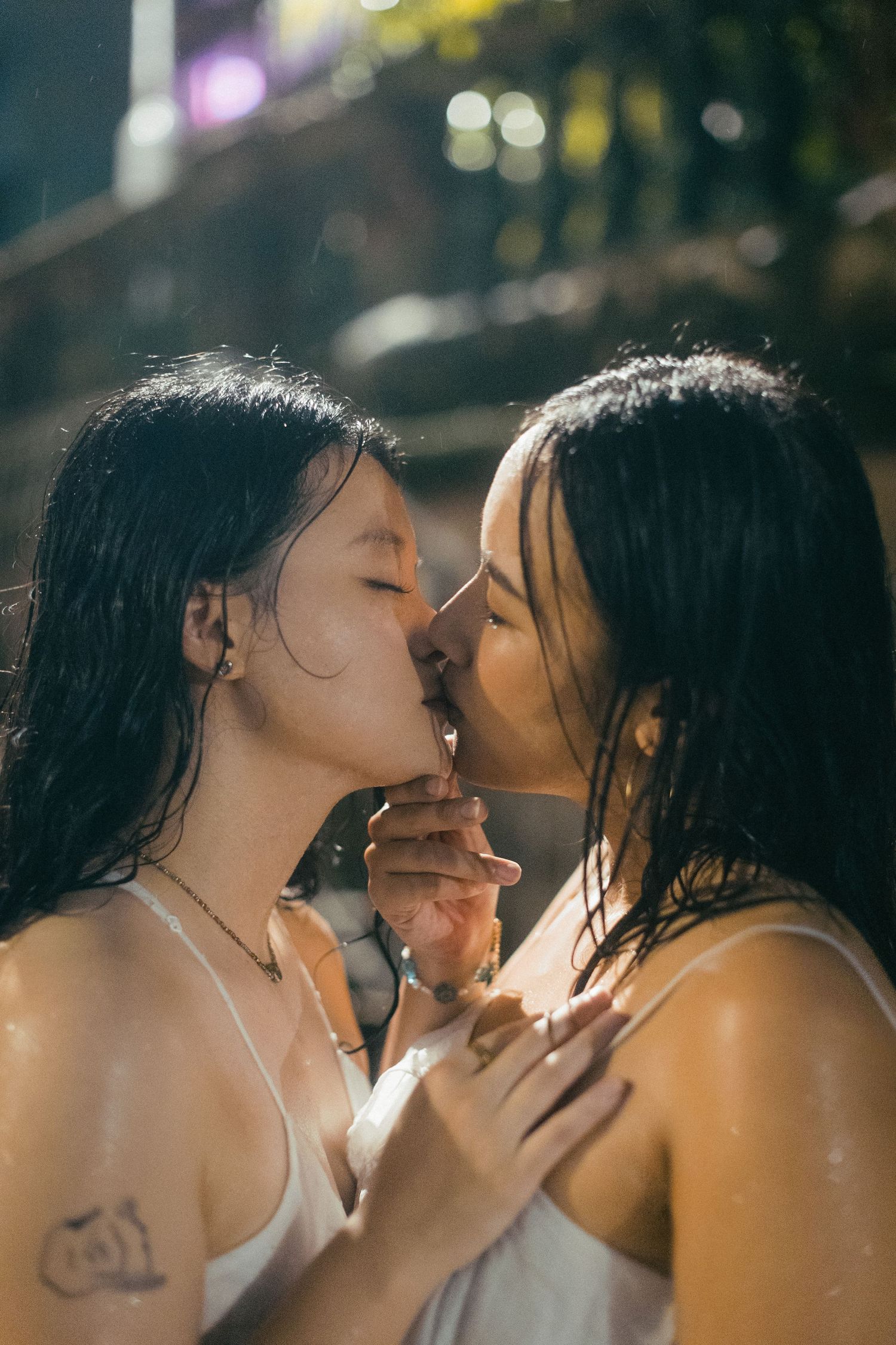 Two wet young Asian women kissing in the rain at night outdoors.